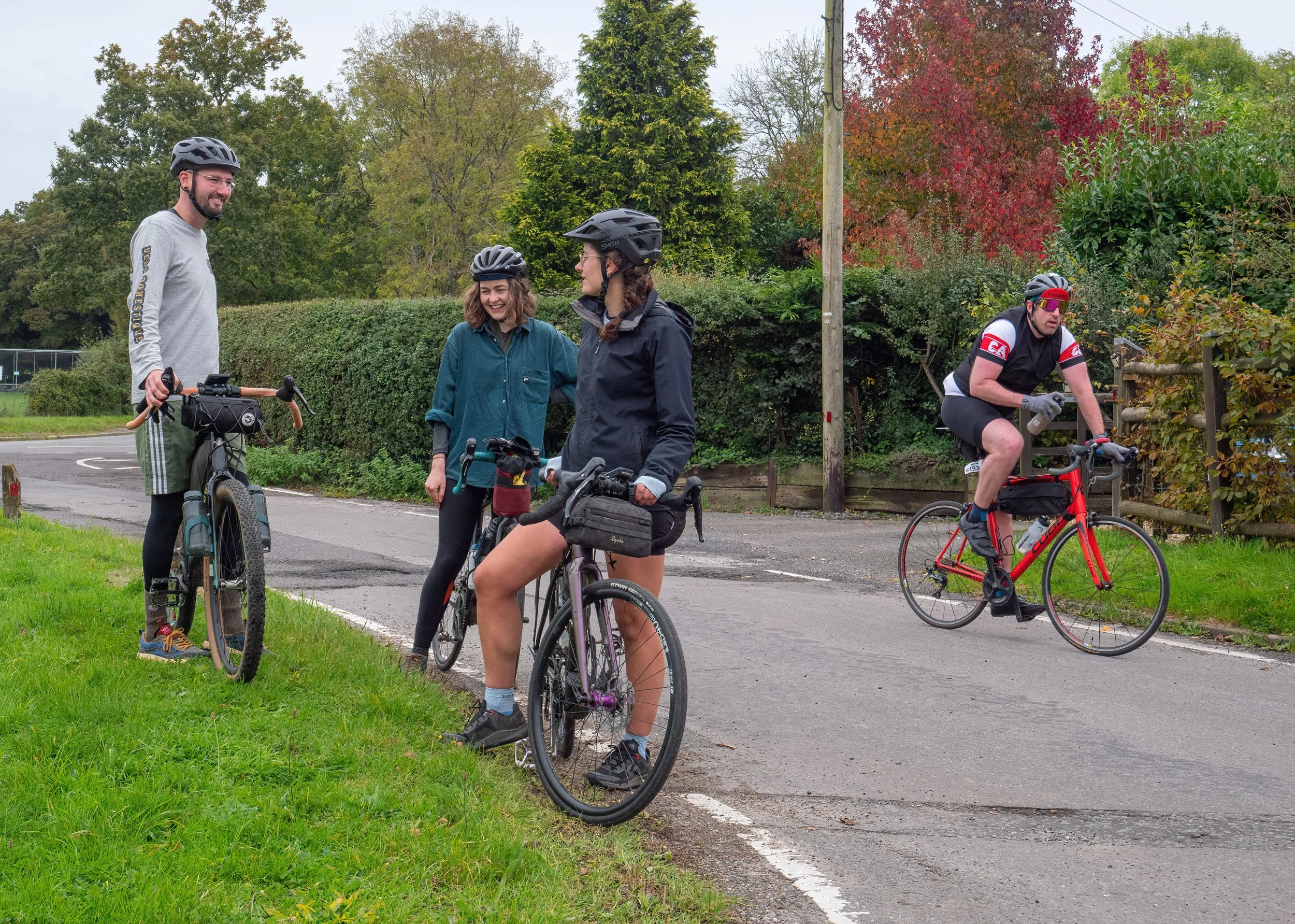 Four people with bicycles conversing on a rural road surrounded by trees displaying fall colors. One person is wearing a gray long-sleeve shirt and helmet, another a teal jacket, and two others are in cycling gear, one with red sunglasses and the oth