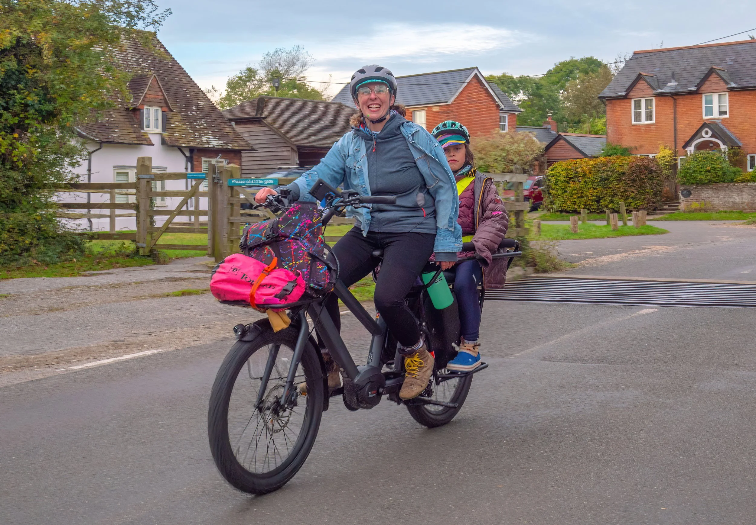 Two women riding a bicycle together along a suburban street with houses and trees in the background, wearing helmets and outdoor clothing.