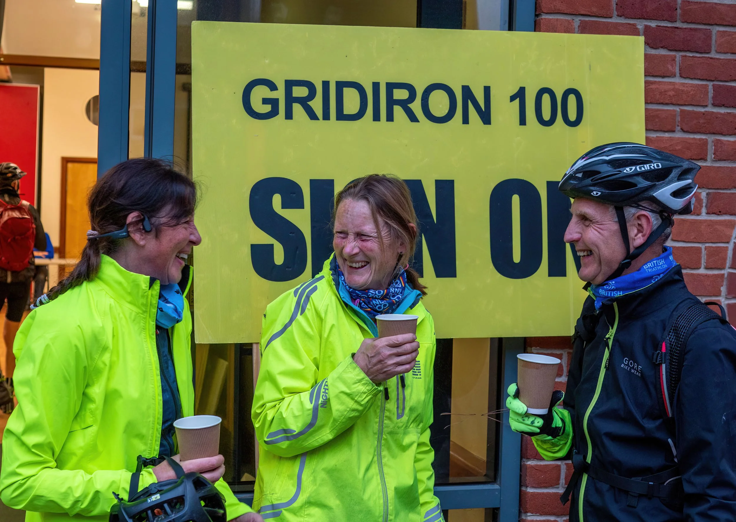 Three smiling people in neon yellow sports jackets standing in front of a yellow sign that reads 'GRIDIRON 100' and 'SUNDAY.' They are holding paper coffee cups and are wearing cycling gear, including a bicycle helmet on one person.