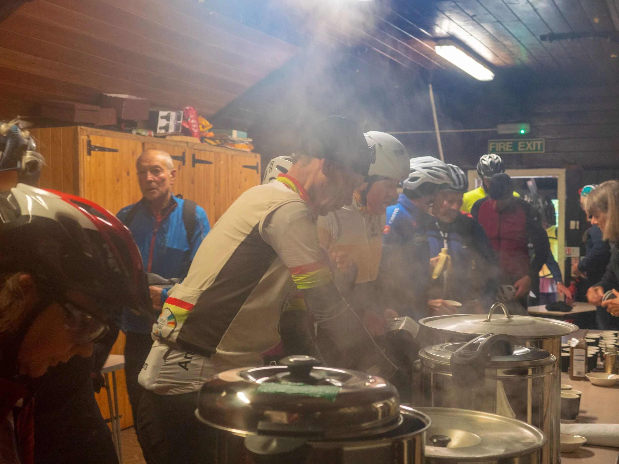 A group of people, some wearing helmets, are gathered around a kitchen counter with large pots, possibly cooking or serving food. The room has wood-paneled walls, shelves with supplies, and a green exit sign. The atmosphere appears busy and communal.