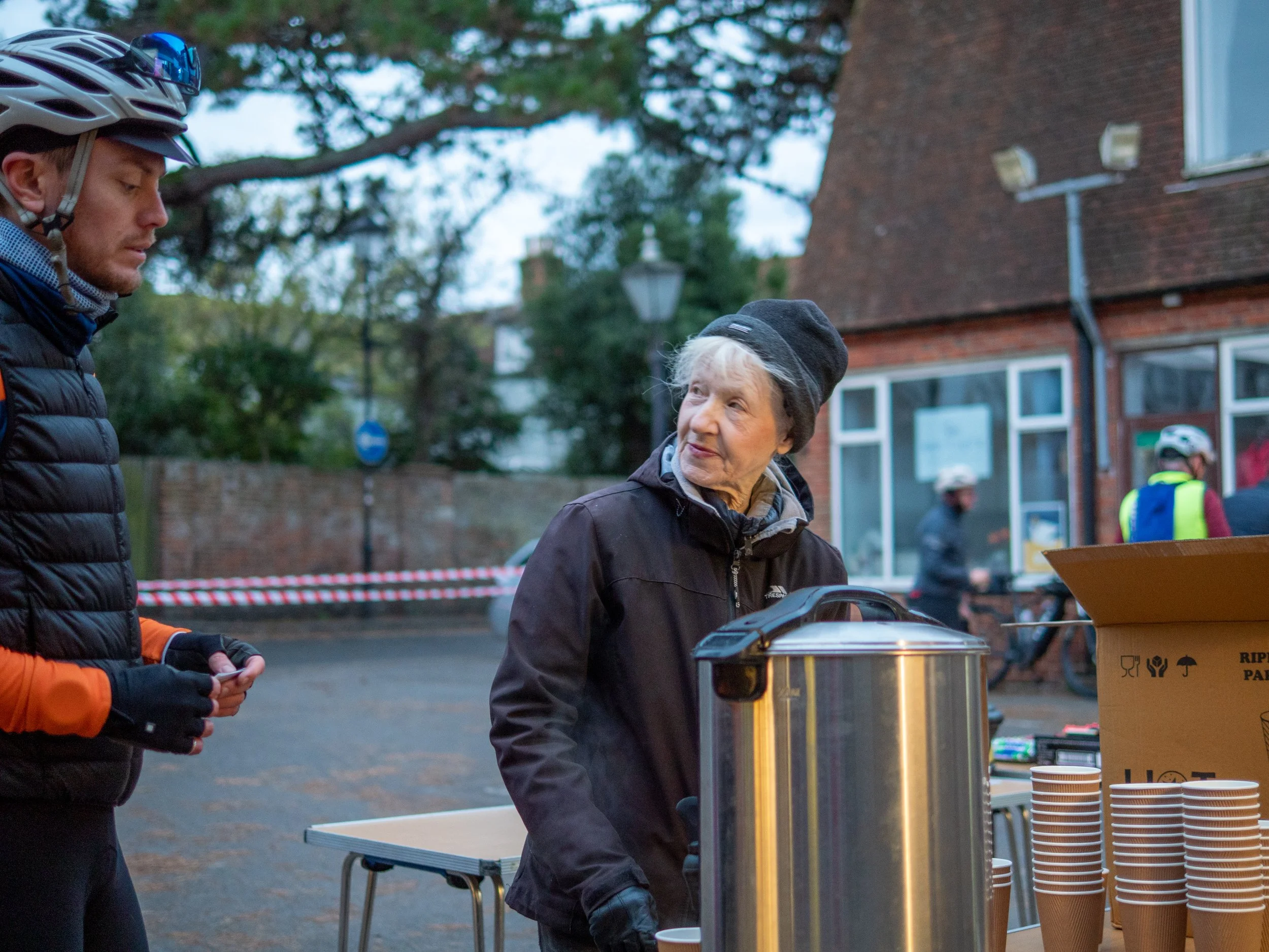 Two men at an outdoor event standing near a table with paper cups and a large container. One man is wearing a black jacket and hat, the other is in a black puffer jacket with a helmet, and they are engaged in conversation. There are other people in t