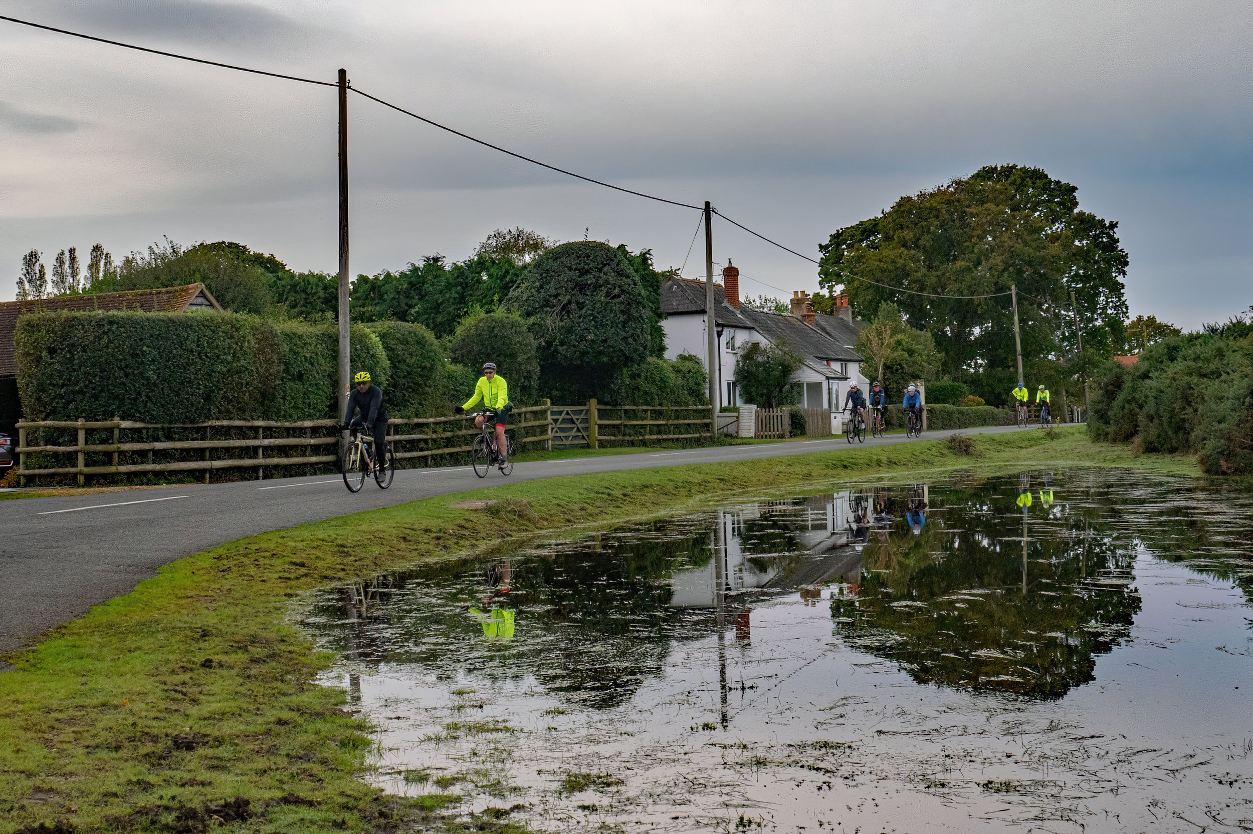 Group of cyclists riding along a rural road beside a flooded area with their reflection visible in the water, under a cloudy sky.