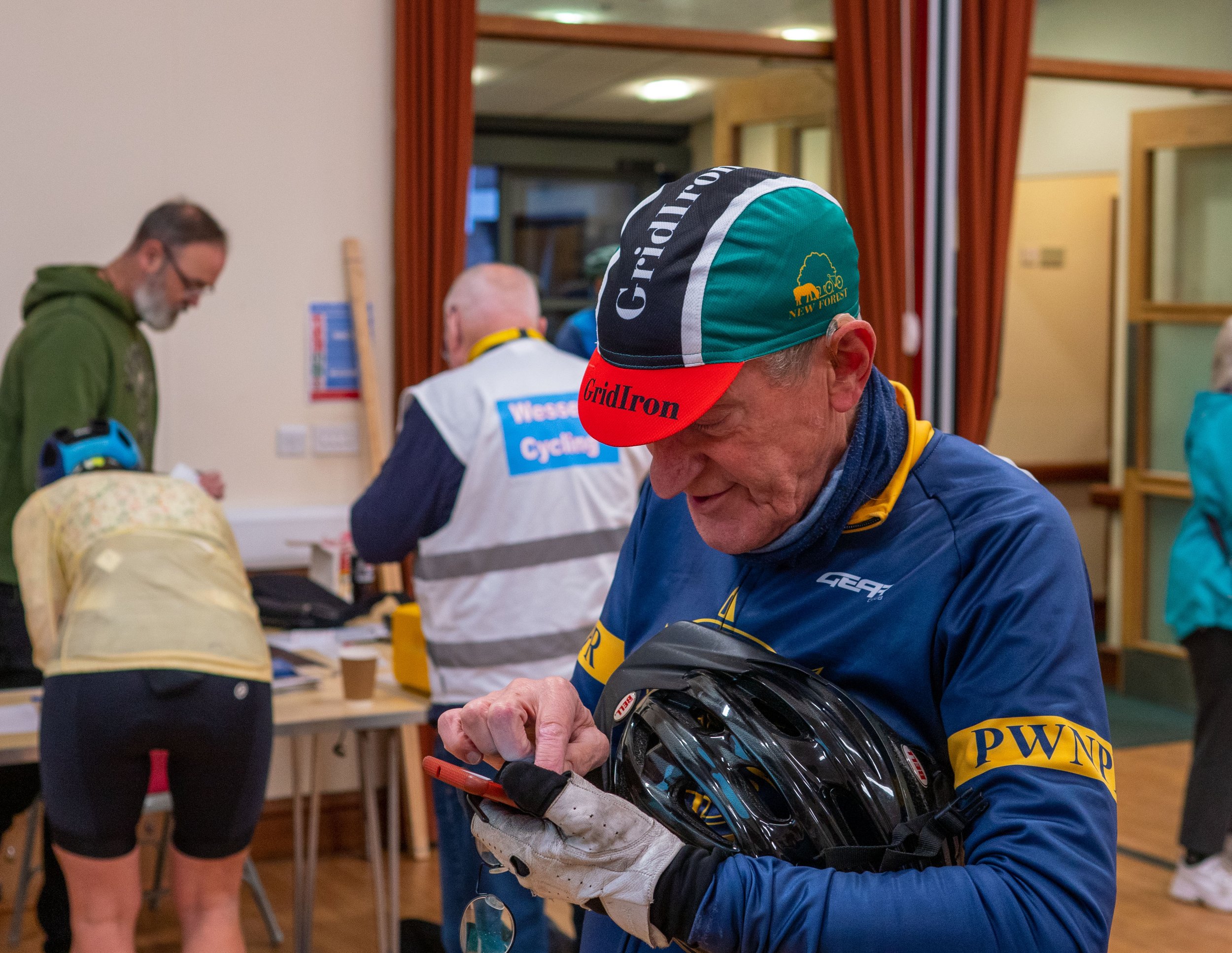 Man in blue jacket and a cycling helmet signing a document, with others in the background in a room with wooden accents and red curtains.