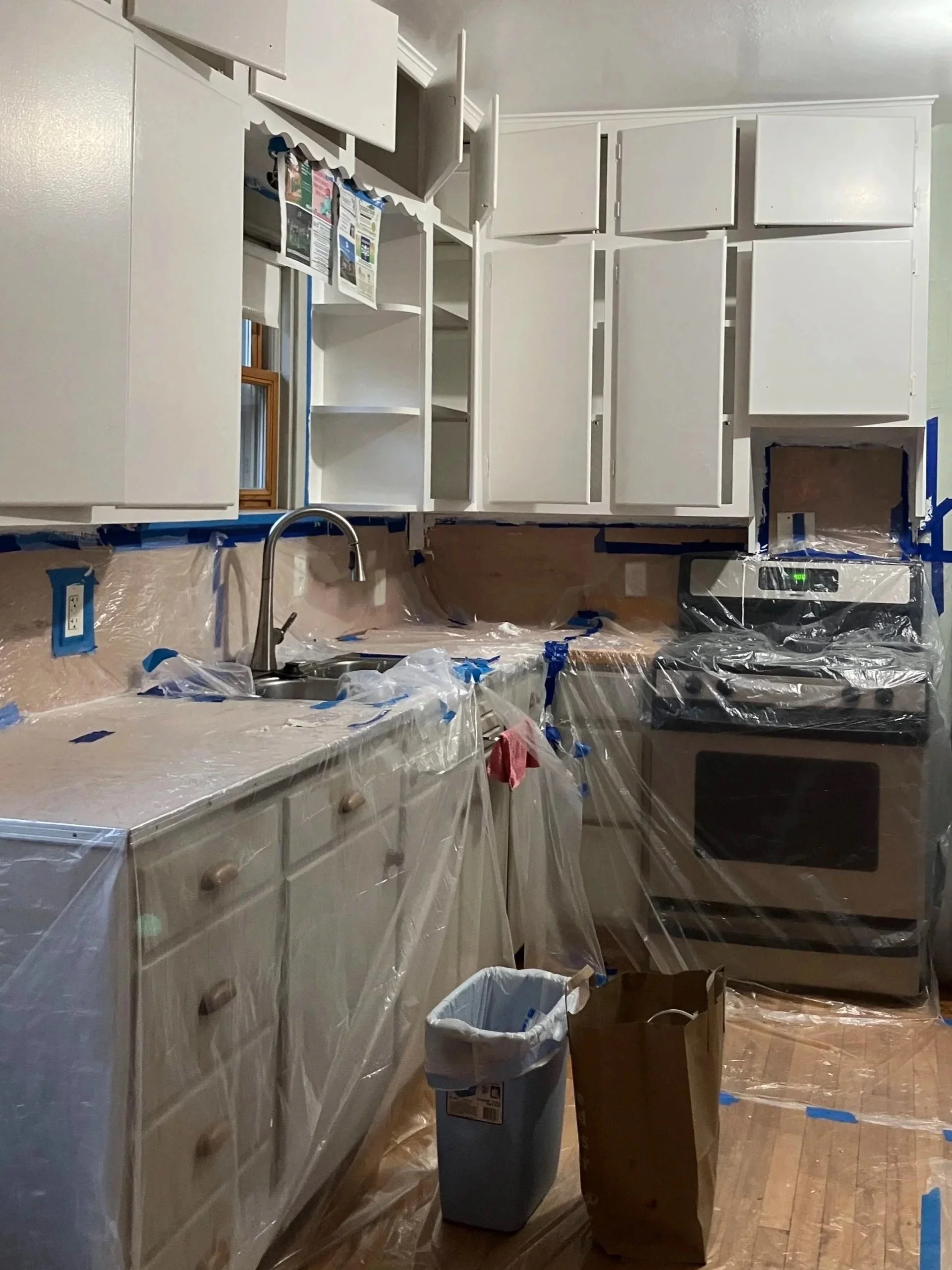 Kitchen under renovation with white cabinets, covered with plastic wrap and blue painter's tape, a faucet over the sink, a stove covered with plastic, and a trash can and paper bag on the floor.