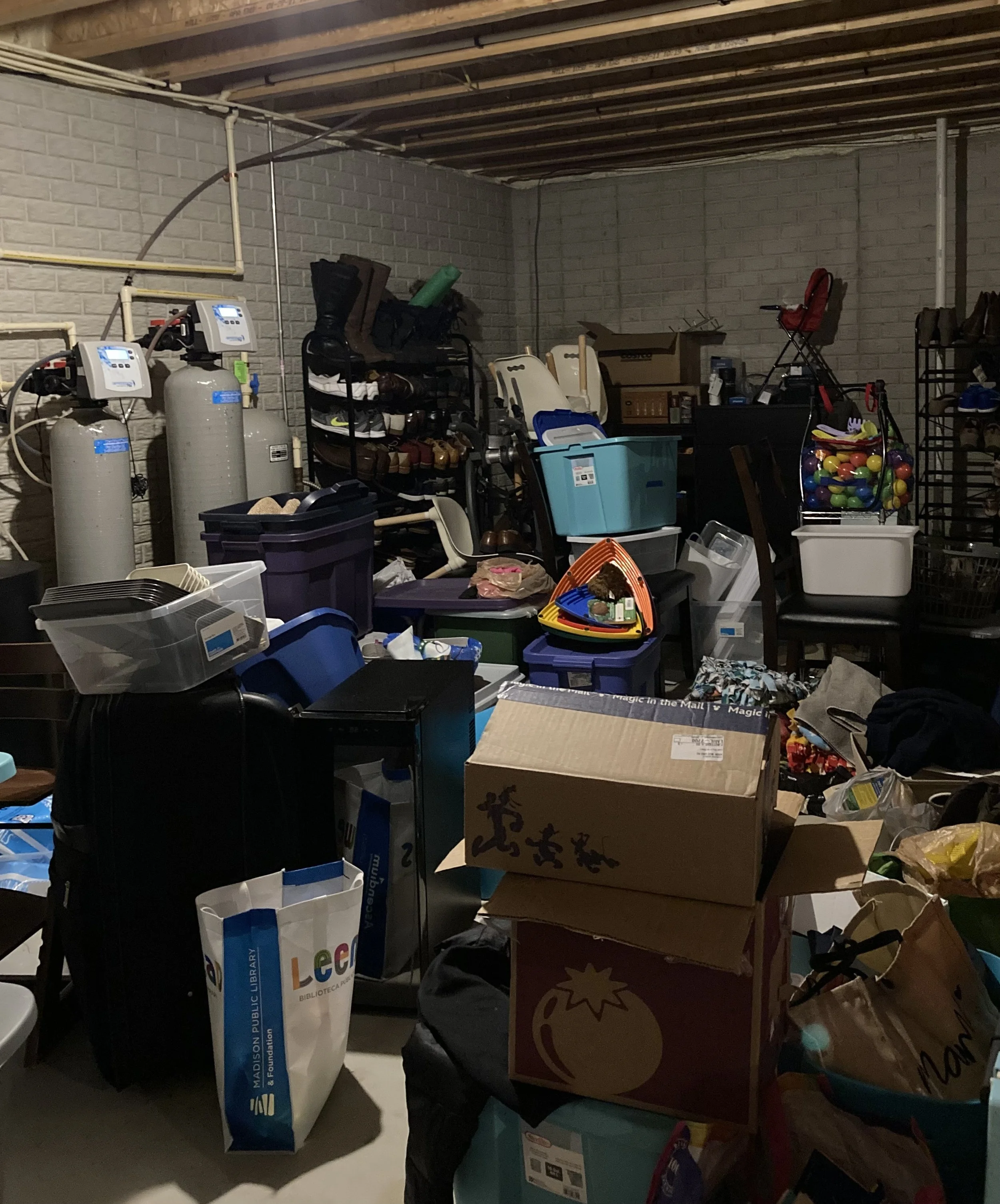 This is a before photo of a basement. There are dozens of shoes, a set of home repair items like lightbulbs, kids books, furniture, and some empty boxes.