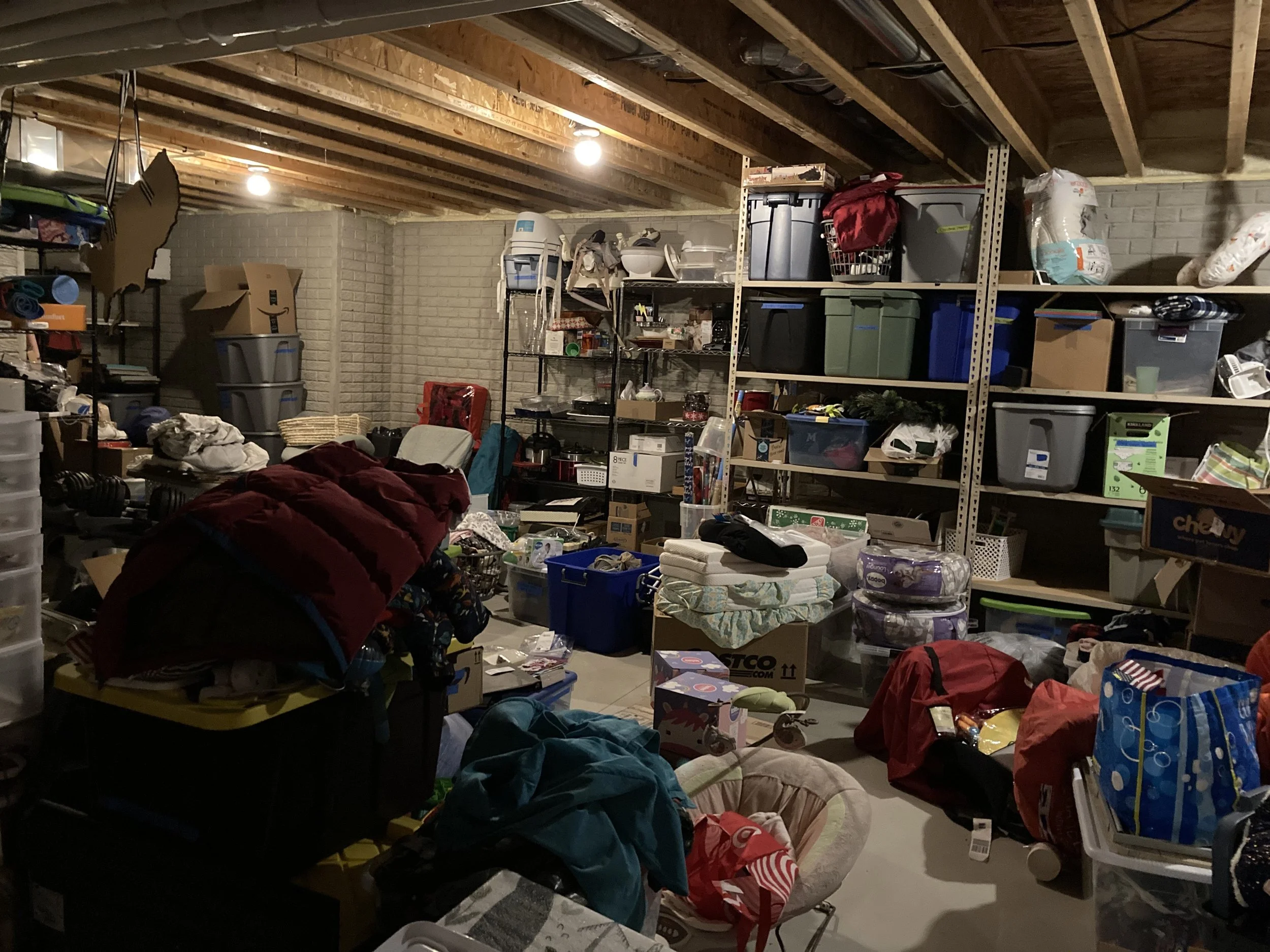 This is a before photo of a basement. Supplies include holiday decor, special cooking equipment, crafting projects, guest room linens, new baby supplies, memory boxes from the previous move, and boxes of professional clothes from a previous job.