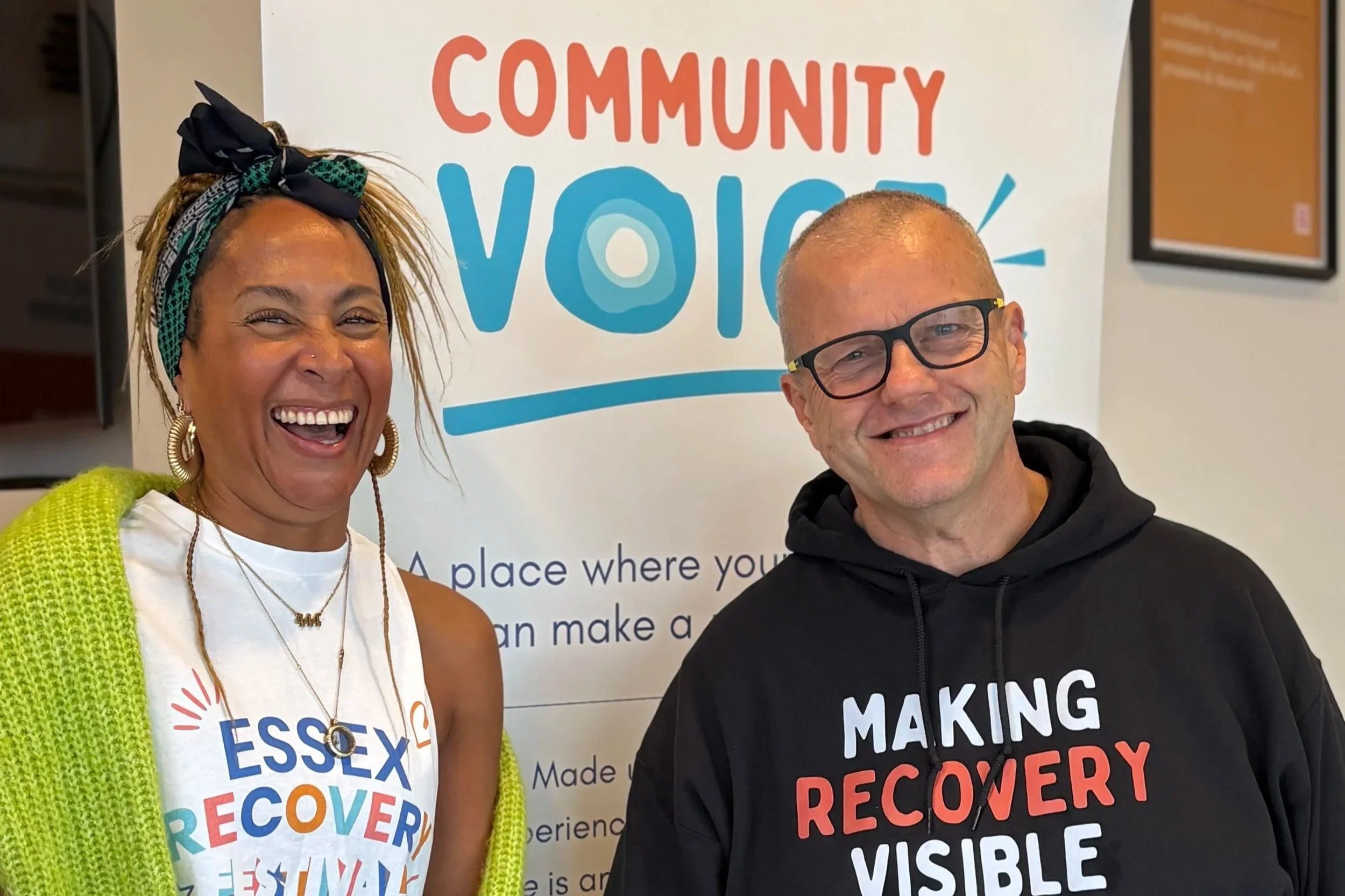 Two community members smile and hold hands in front of a Community Voice banner. One wears an Essex Recovery Festival T-shirt and the other a “Making Recovery Visible” hoodie.