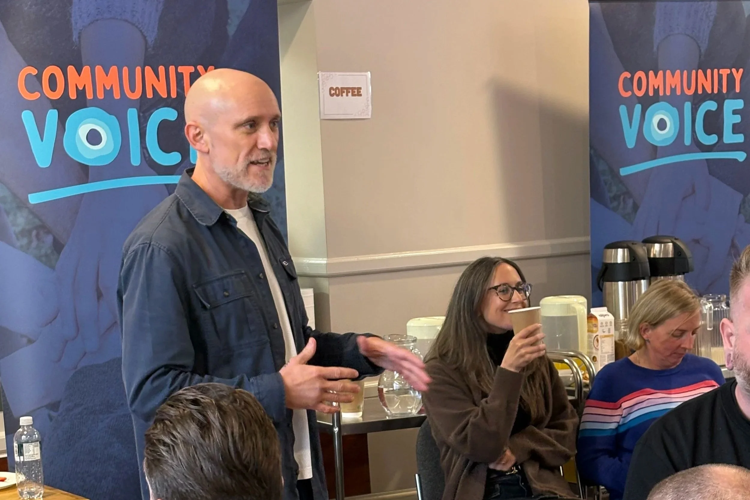 Laurence addresses the room in front of Community Voice banners while people listen and drink coffee.