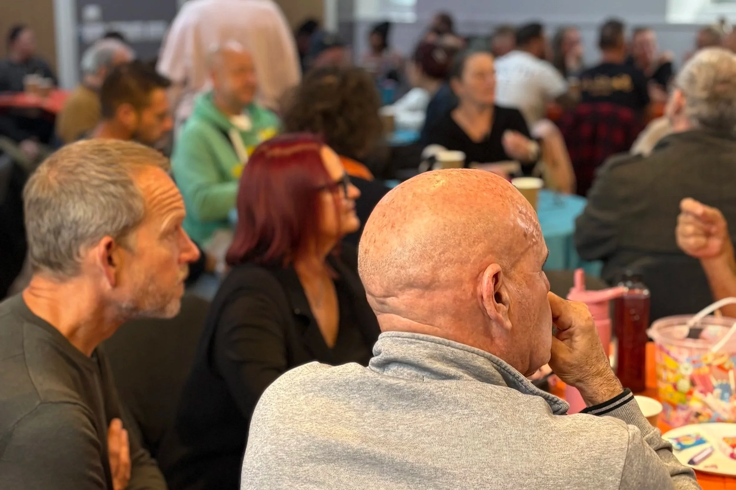 A full room of community members sit around tables listening and chatting at Community Voice in Rayleigh.