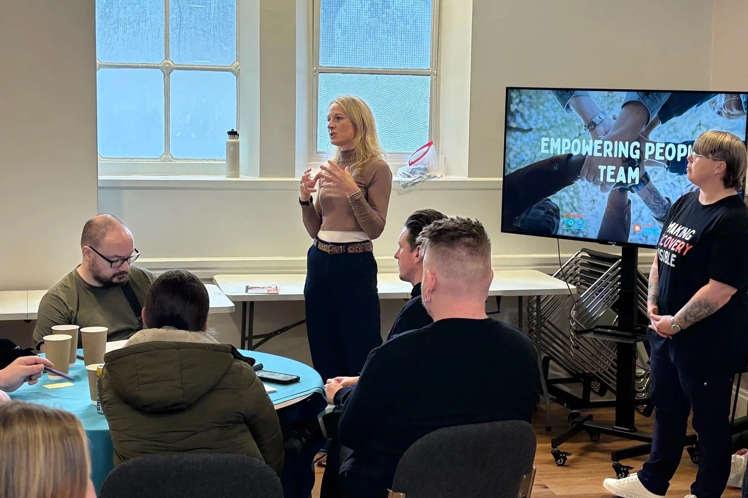 A member of the Empowering People team speaks to a group seated around tables. A colleague in a “Making Recovery Visible” T-shirt stands beside a screen showing the team’s slid