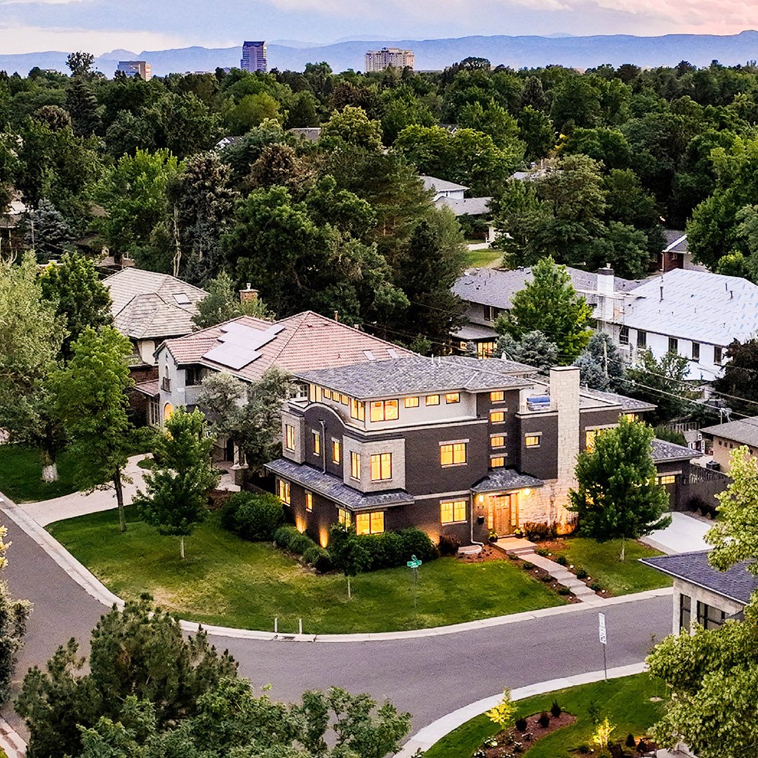A large modern house with illuminated windows, surrounded by green trees and a well-maintained lawn. The house is situated on a curved street in a suburban neighborhood with other houses and taller buildings in the background.