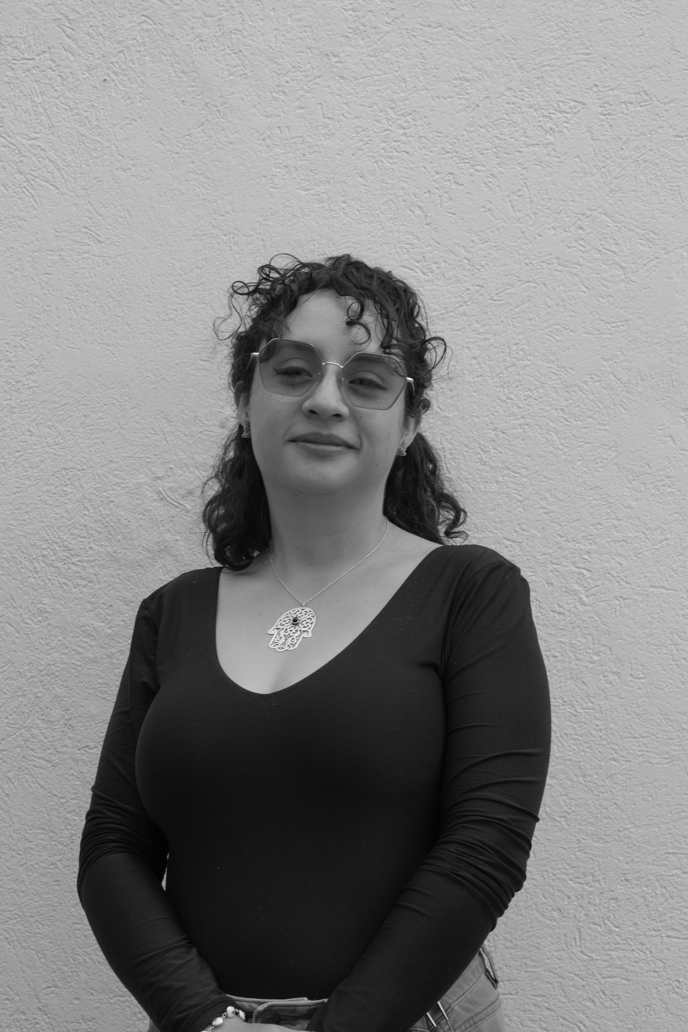 Retrato de mujer con cabello rizado y gafas grandes de sol, usando una blusa negra con escote y un collar decorativo, posando contra una pared de textura rugosa.