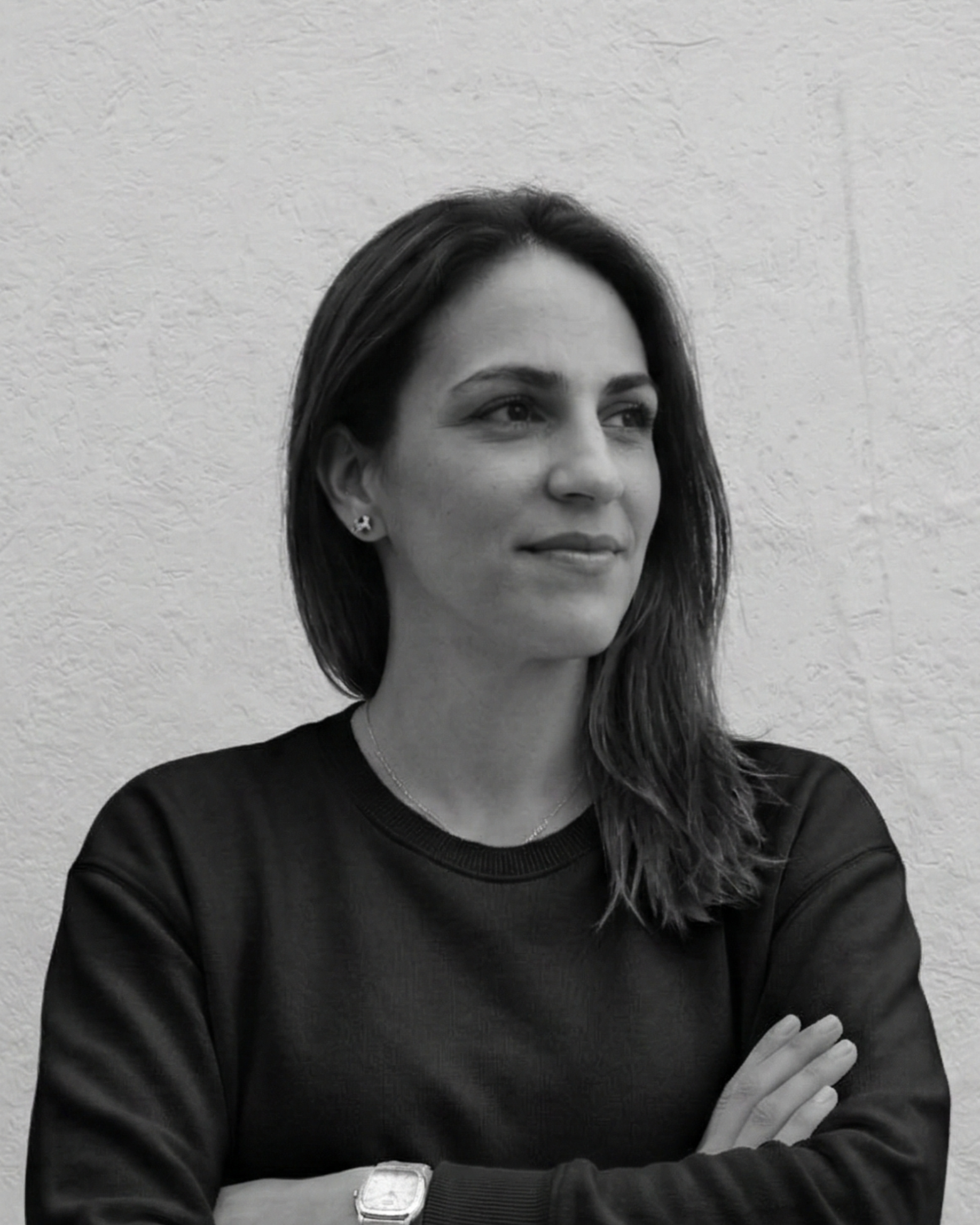 Retrato en blanco y negro de una mujer con cabello largo y oscuro, usando una camiseta negra y con la mano sobre el brazo. La mujer tiene pendientes de estrella y una pulsera en la muñeca, vista de perfil con expresión neutral, frente a una pared de textura rugosa.