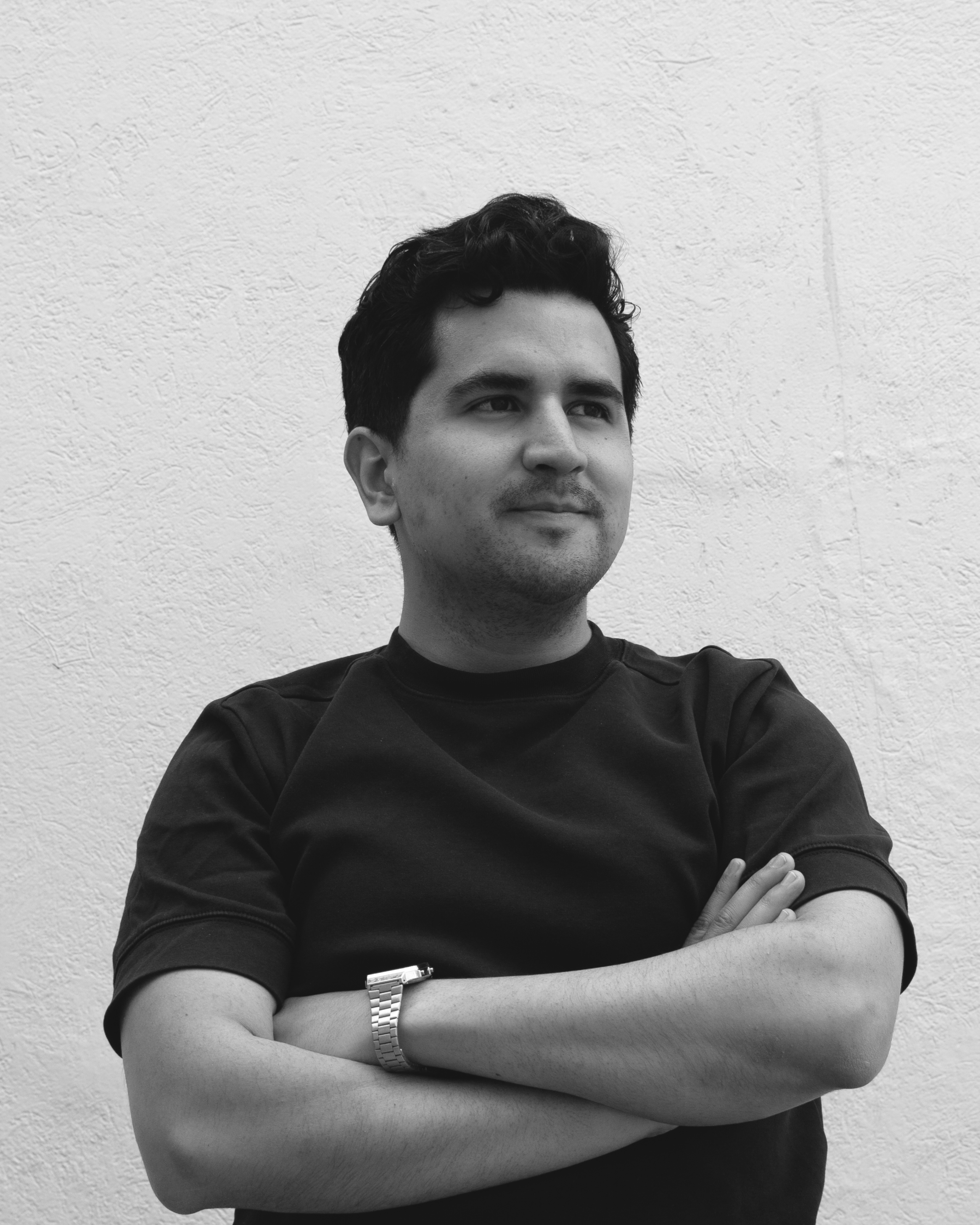 Hombre joven con cabello oscuro, barba ligera, cruzado de brazos, usando camiseta negra y relojplateado, de pie frente a pared clara con textura.