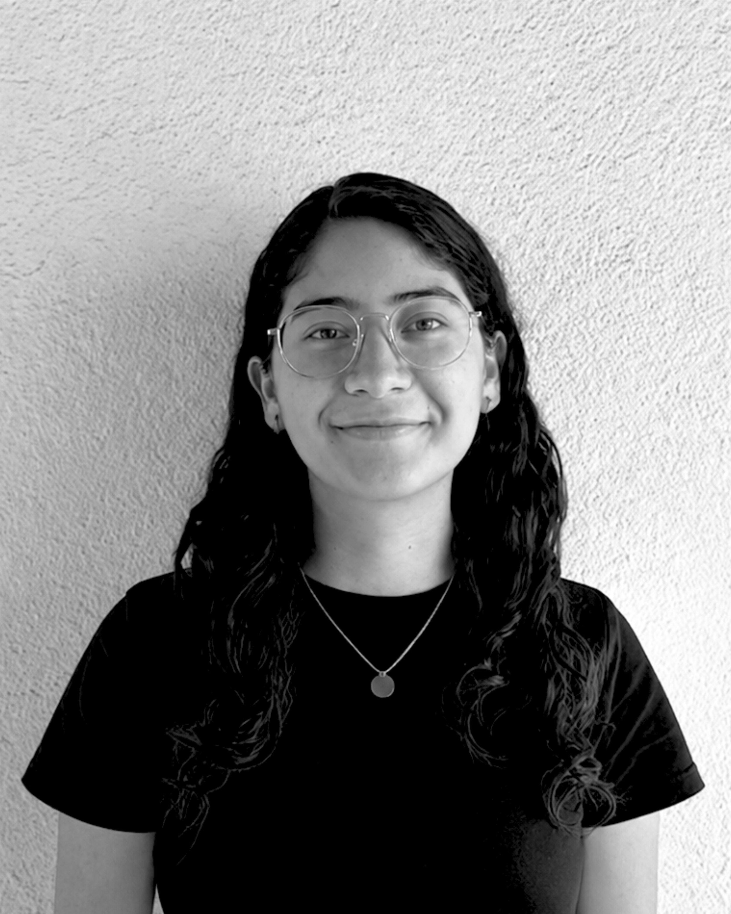 Retrato en blanco y negro de una joven mujer con gafas, sonriendo, con cabello rizado y camiseta negra, frente a una pared de textura rugosa.
