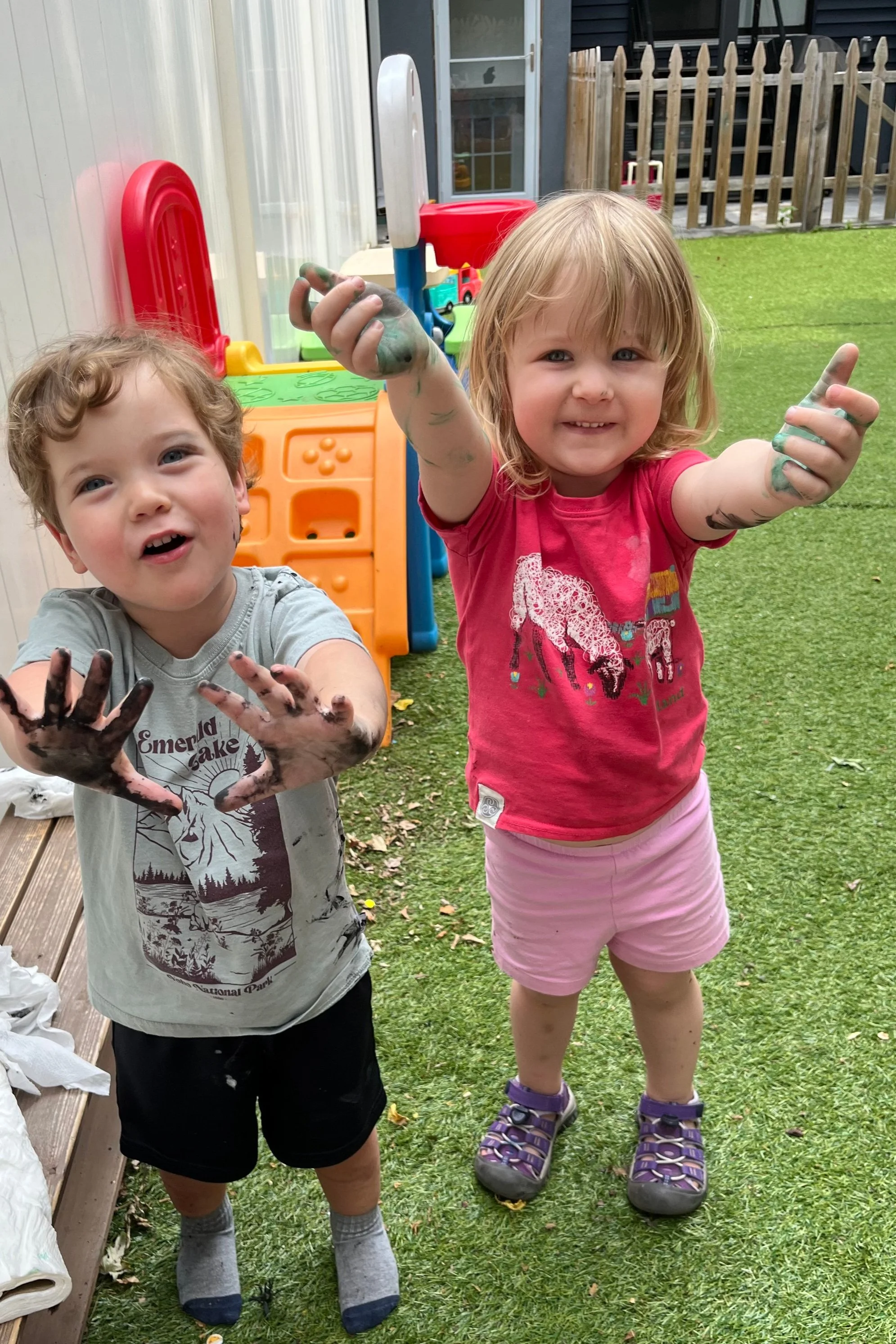 Two young children, a boy and a girl, playing outside with messy hands covered in black and green paint, smiling and showing their paint-covered hands towards the camera on a grassy yard.