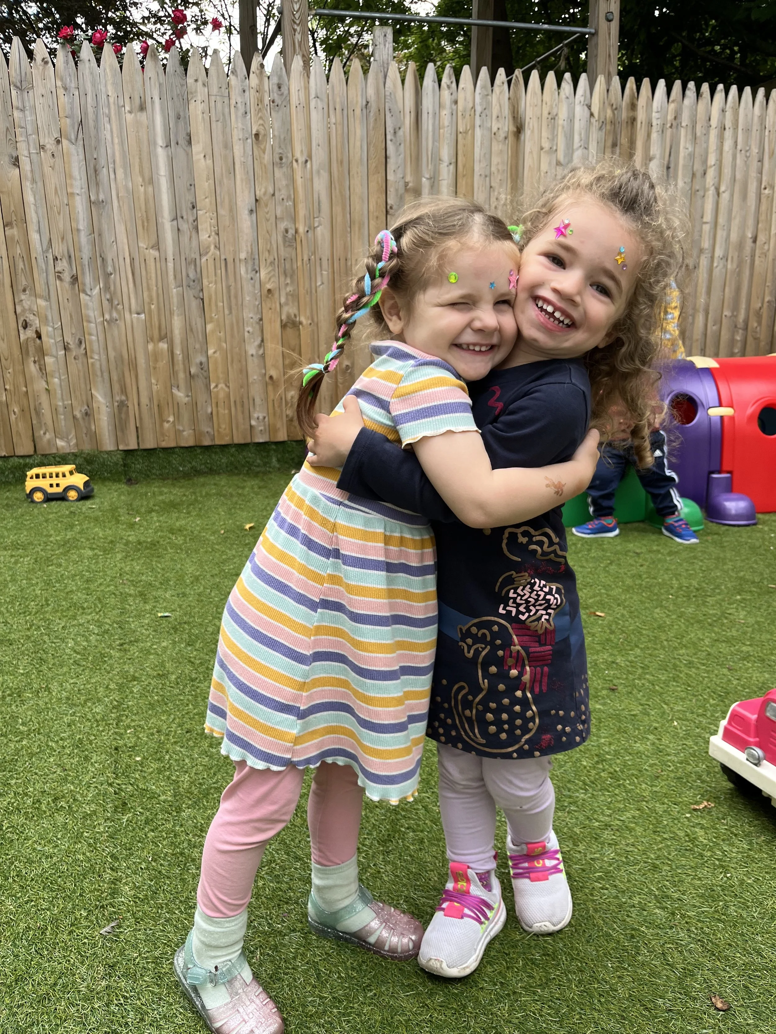 Two young girls hugging and smiling in a backyard with a wooden fence, colorful toys, and grass.