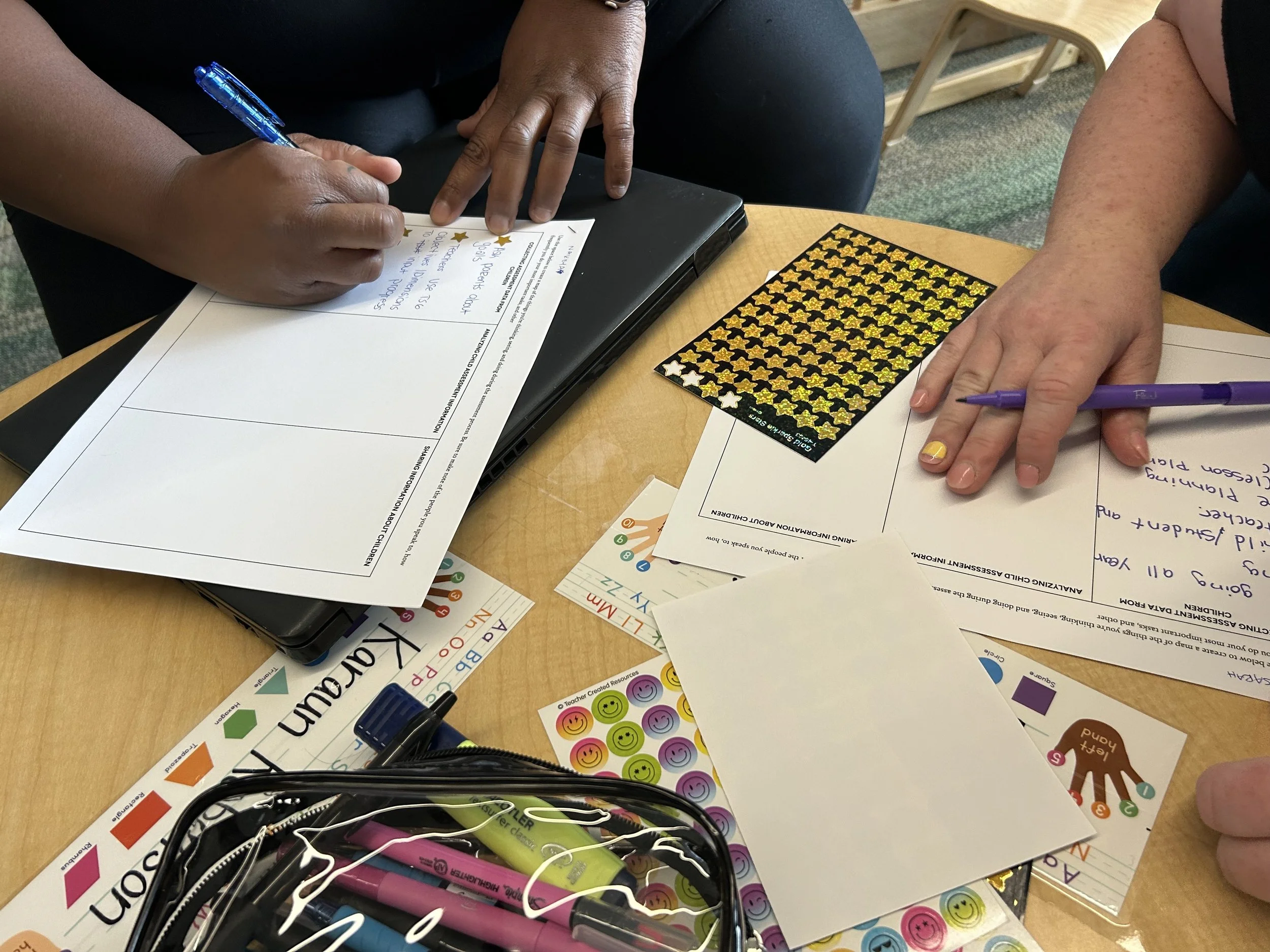 Two people working at a table with papers, stickers, and pens. One person is writing on a worksheet with a blue pen, another is holding a purple pen. There are various colorful stickers, a closed black laptop, and a clear pencil case filled with markers on the table.