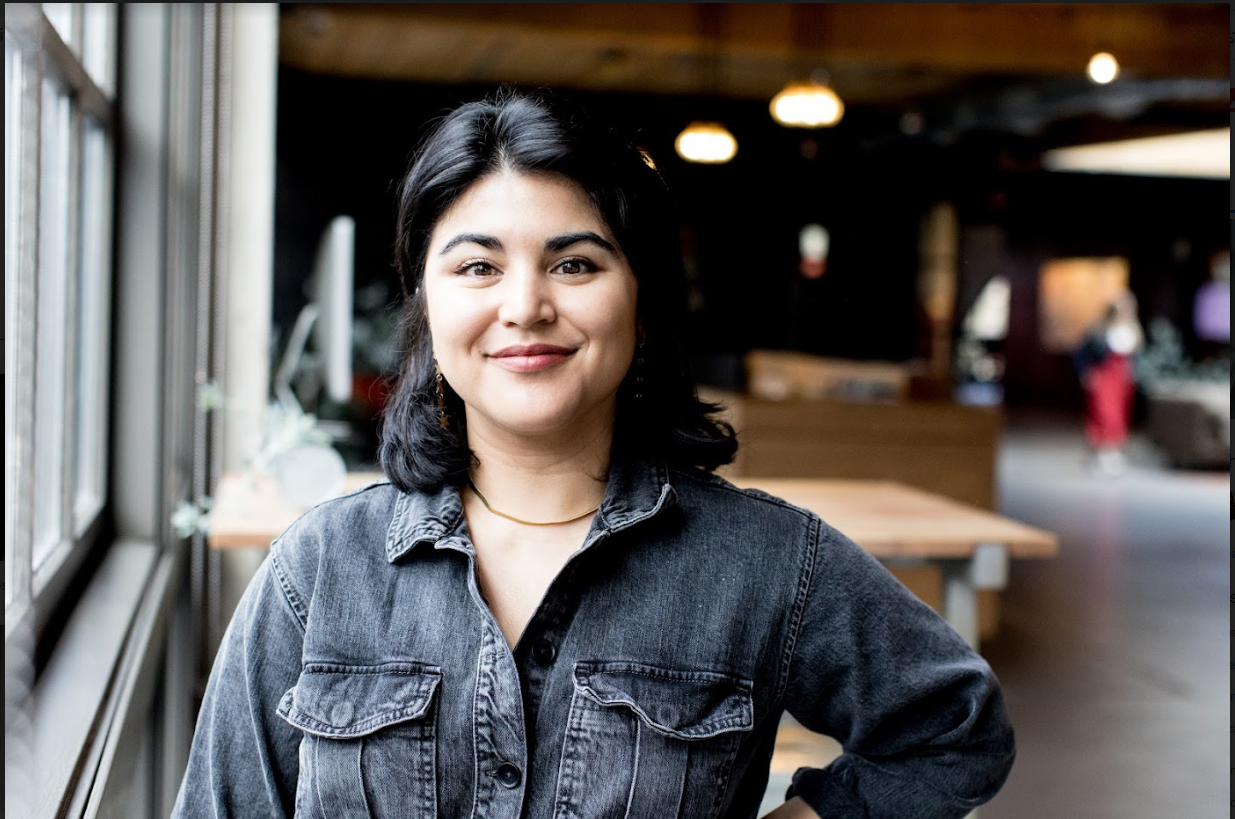A young woman with black hair, wearing a dark denim jacket, smiling while standing inside a cafe near a large window.