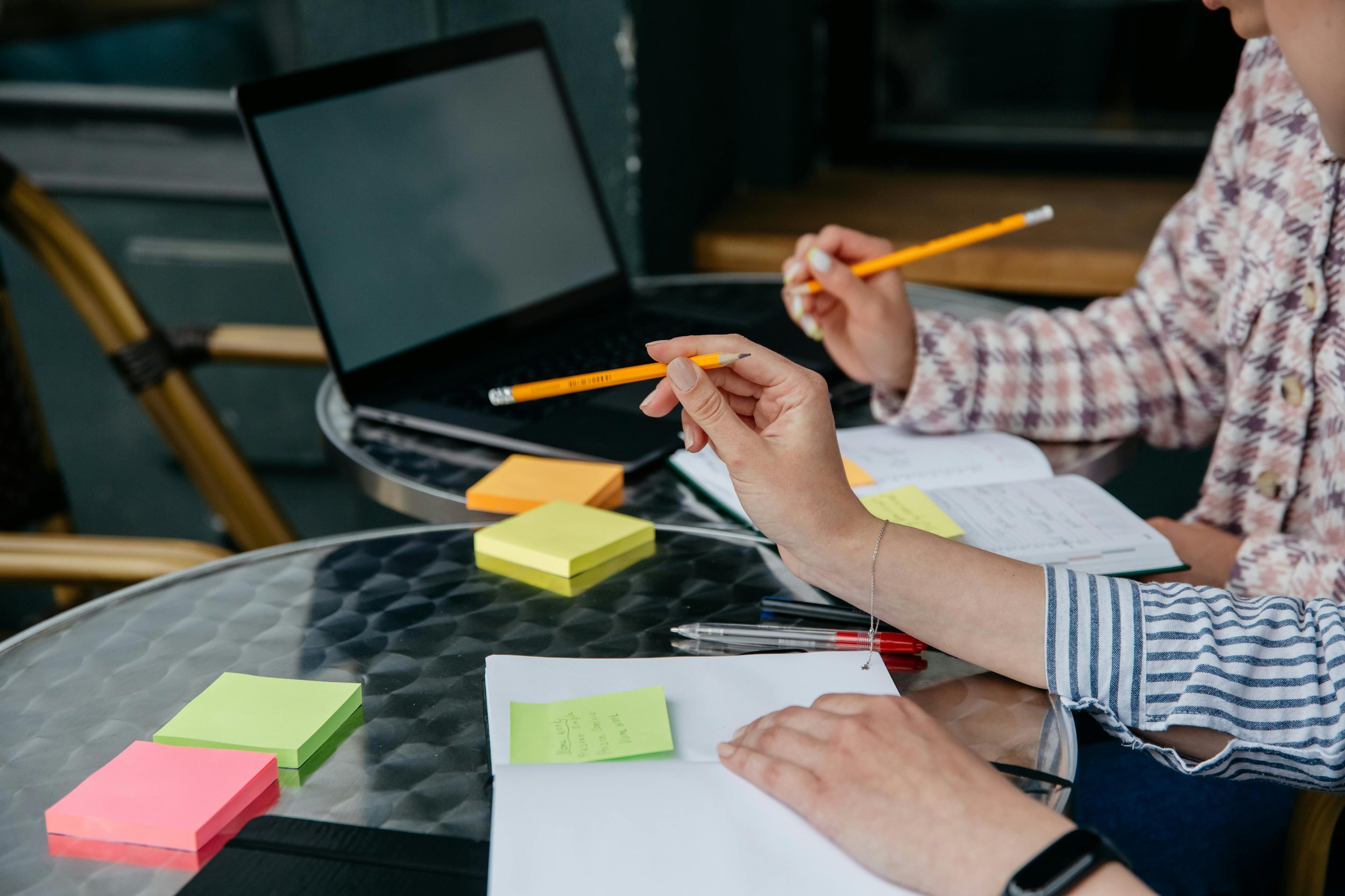 Two people working at a table with sticky notes, pens, a notebook, and a laptop.