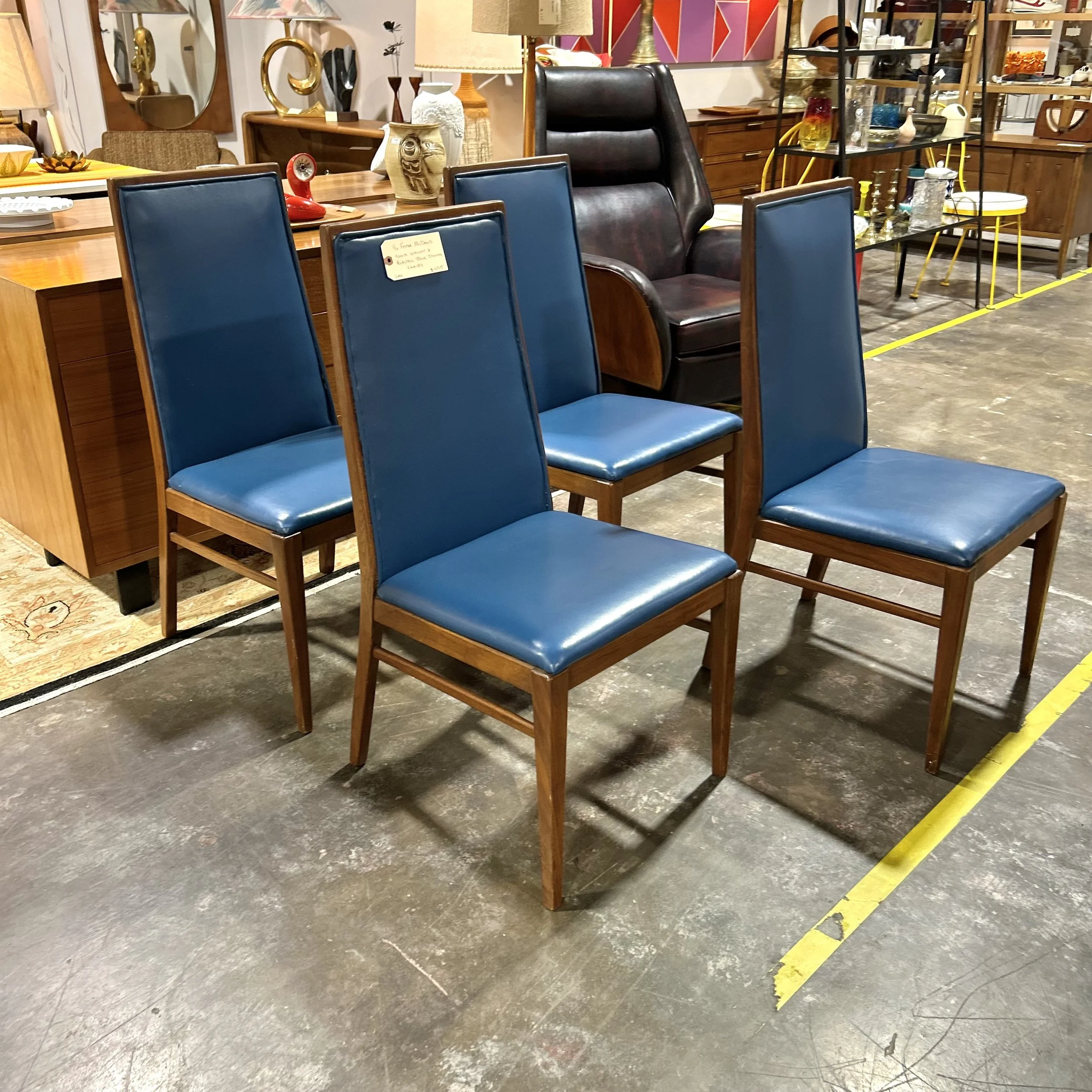 A group of four vintage wooden chairs with blue vinyl seats and backs arranged in a square in a furniture store. Background displays various lamps, decorative items, and furniture pieces.