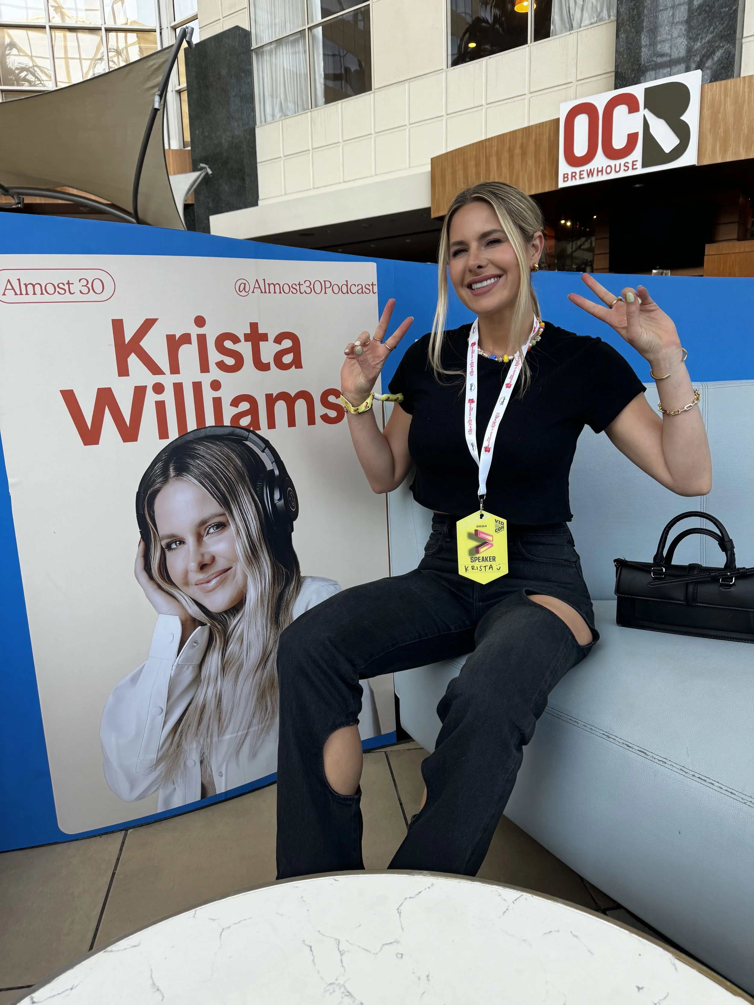 Krista Williams sitting on a couch at a podcast event, smiling and making peace signs with her hands. She is wearing a black t-shirt, black ripped jeans, and colorful jewelry, with a sign hanging around her neck that says "Speaker Krista." There is a large promotional poster with her photo and name next to her, and a sign for OC Brewhouse in the background.