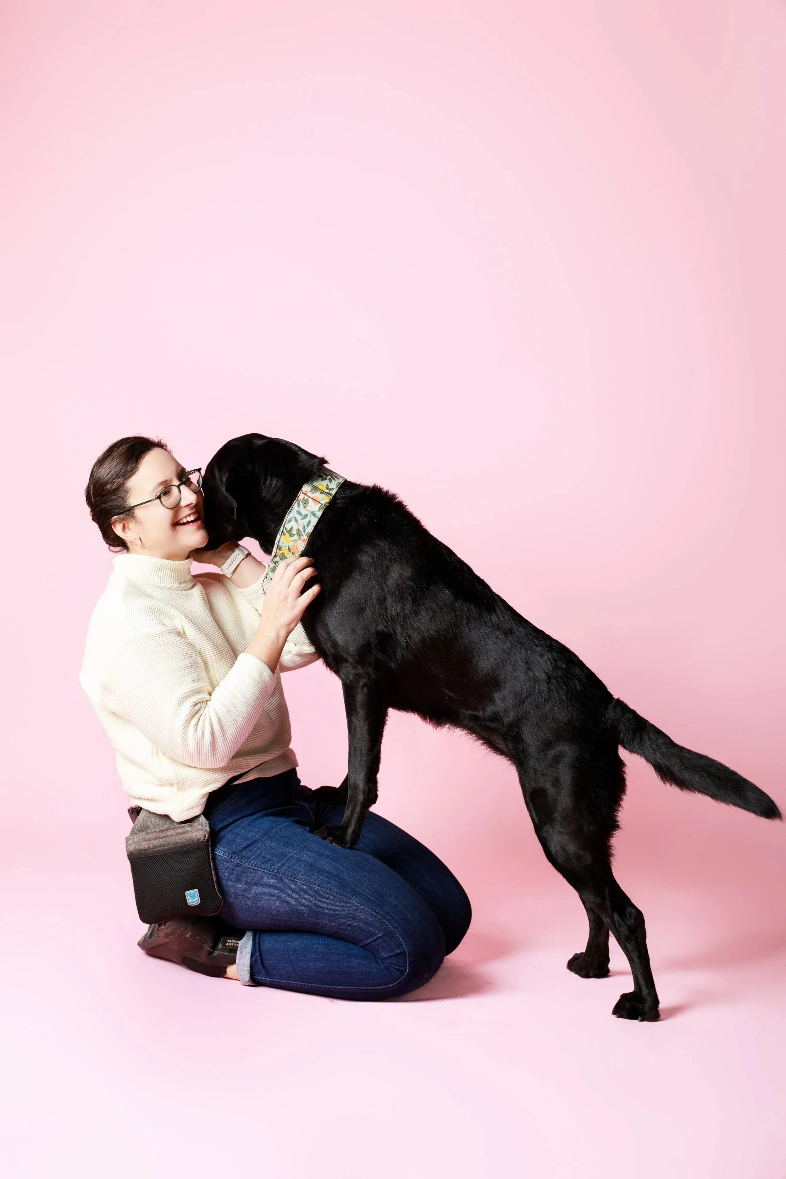 A dog trainer kneels with a black labrador retriever standing on her lap and licking her face