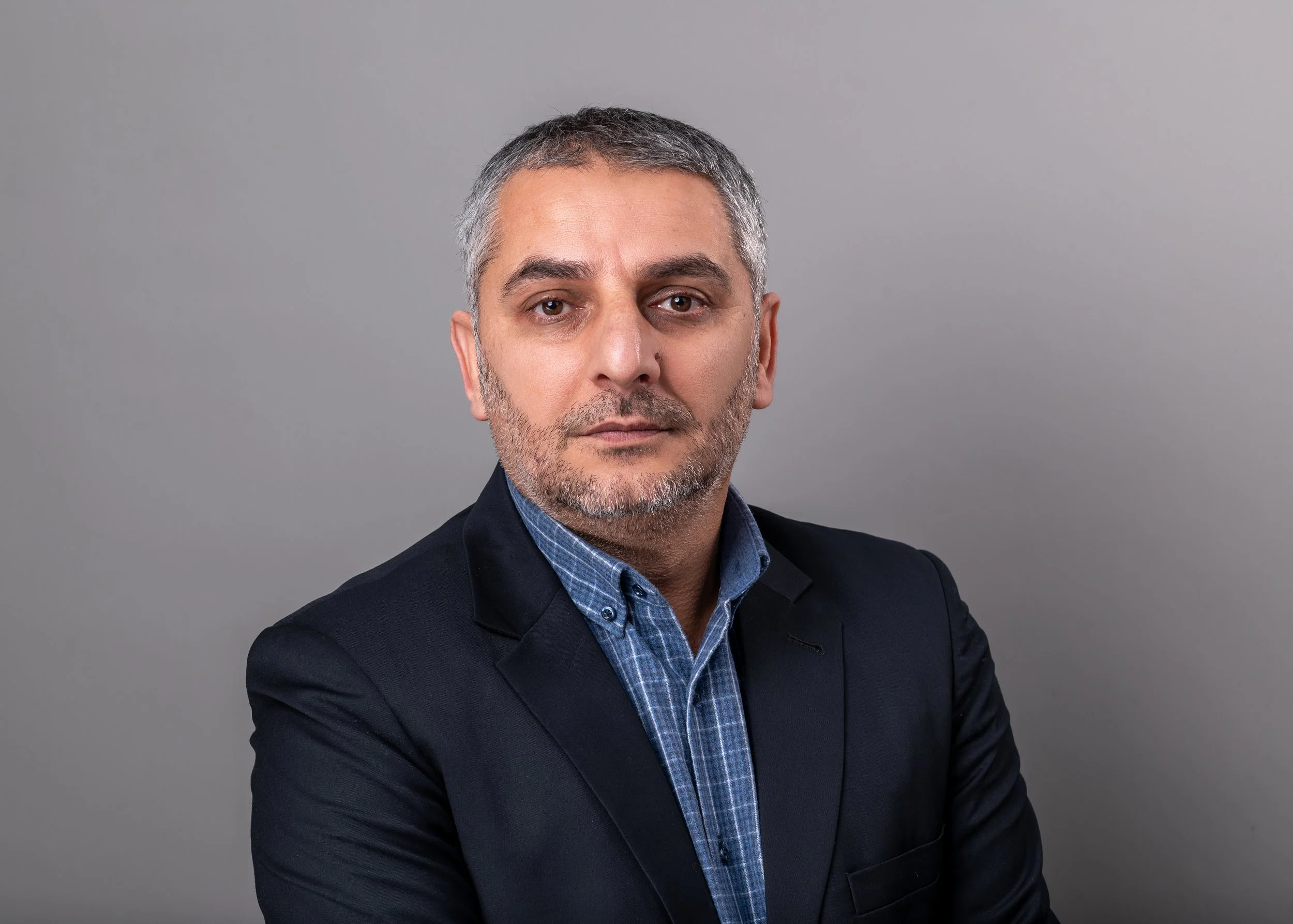 image of male with gray hair and beard, wearing a black blazer with a blue undershirt, with a gray background