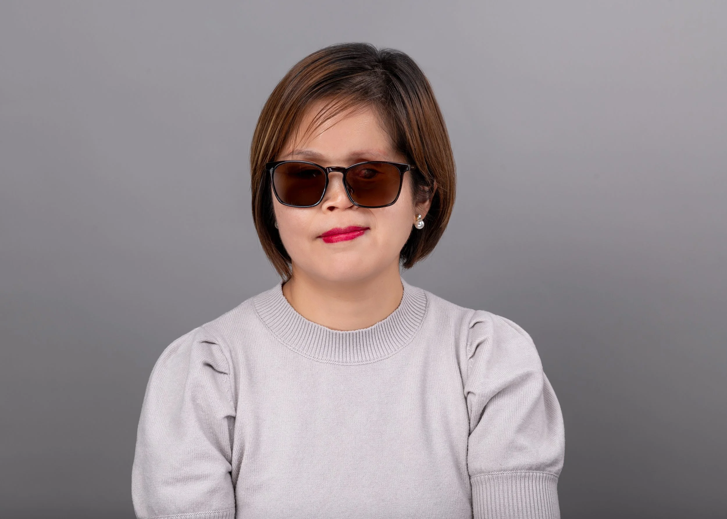 image of female with short brown hair, wearing dark glasses, with a gray shirt, and a gray background