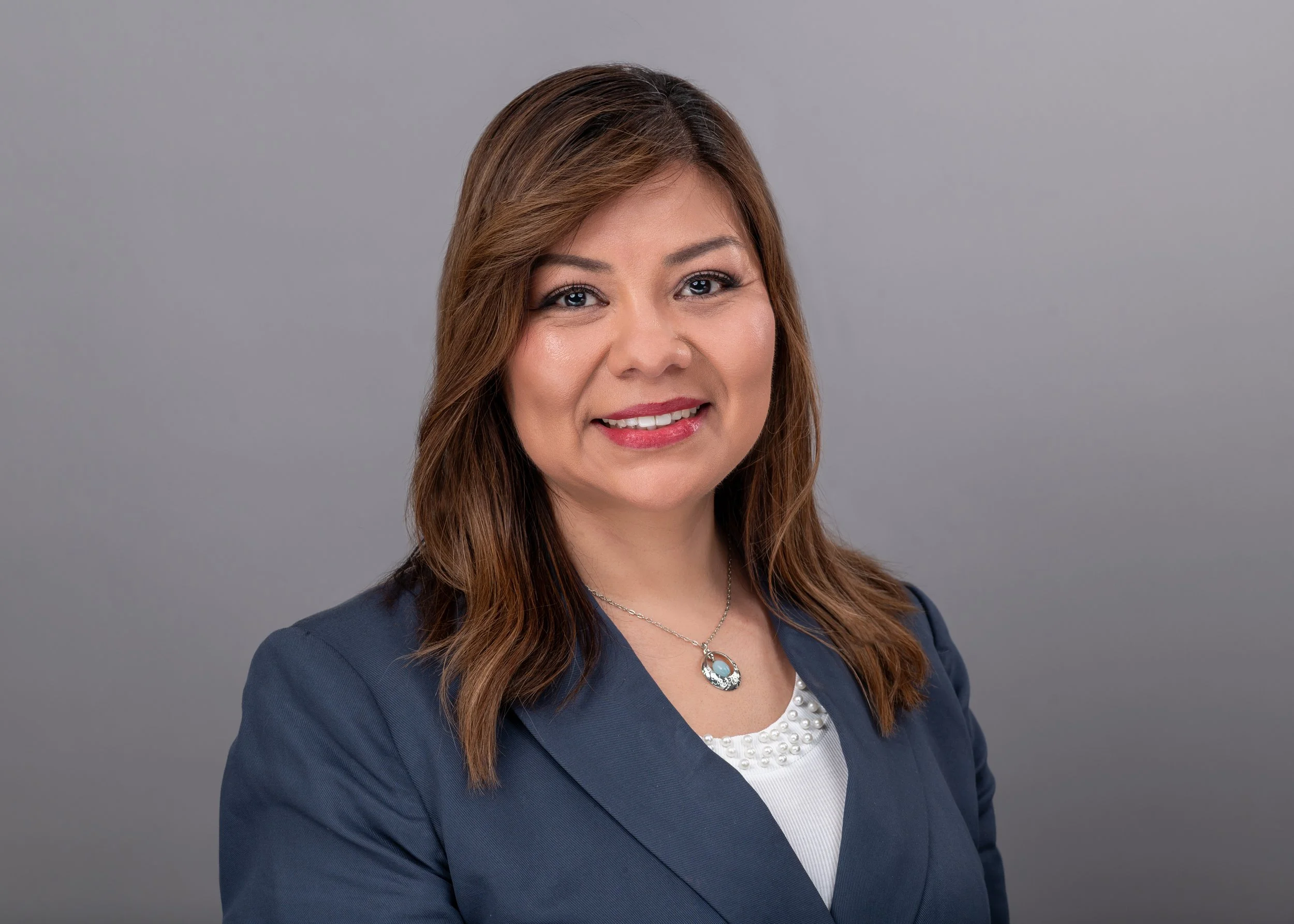 photo of Latina woman with brown hair, wearing a navy blazer and white shirt, with gray background
