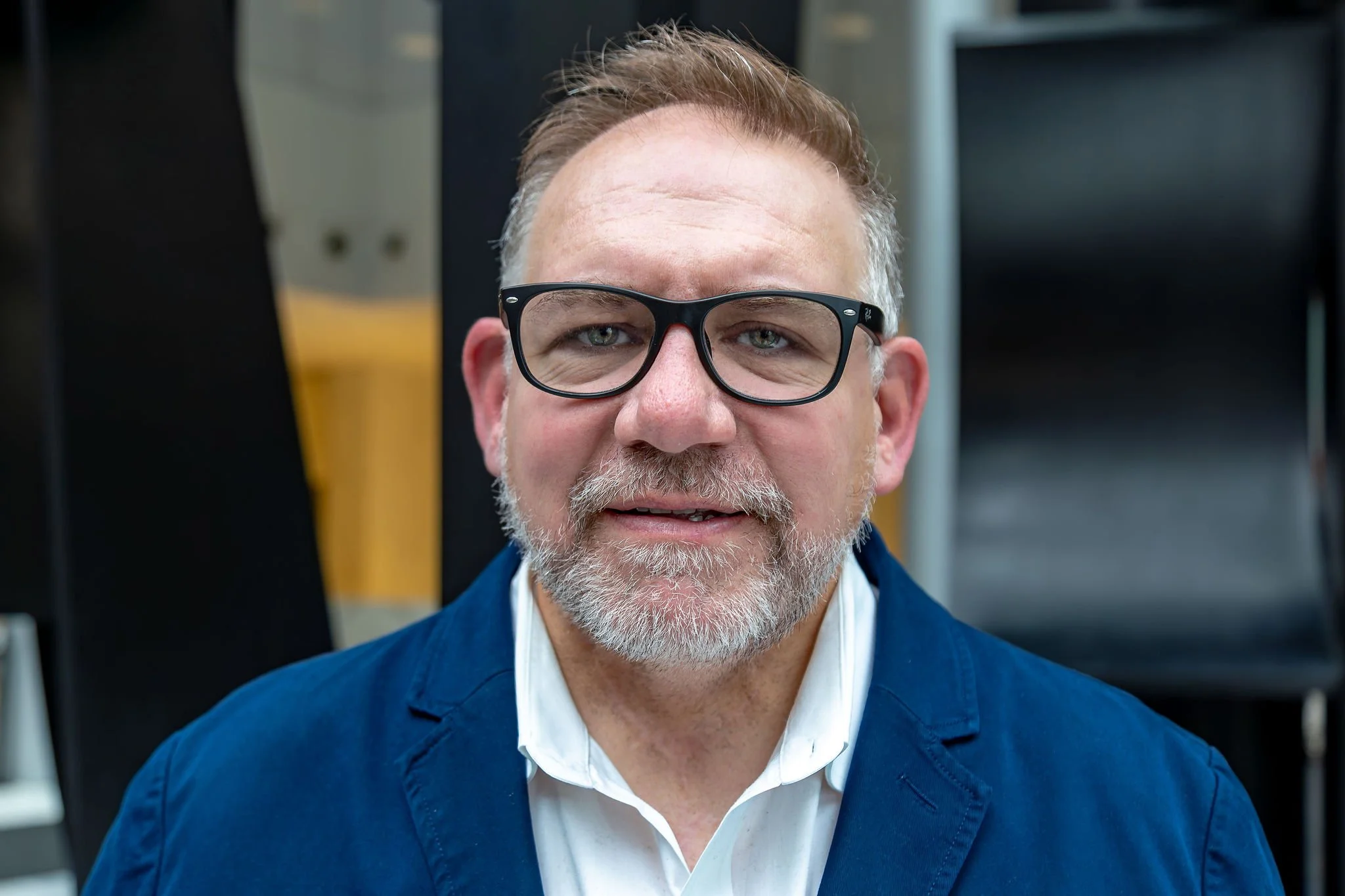 Mark is pictured in an open space with a textured background. He has a slight smile and is wearing black glasses, a navy blue and a white collared shirt.