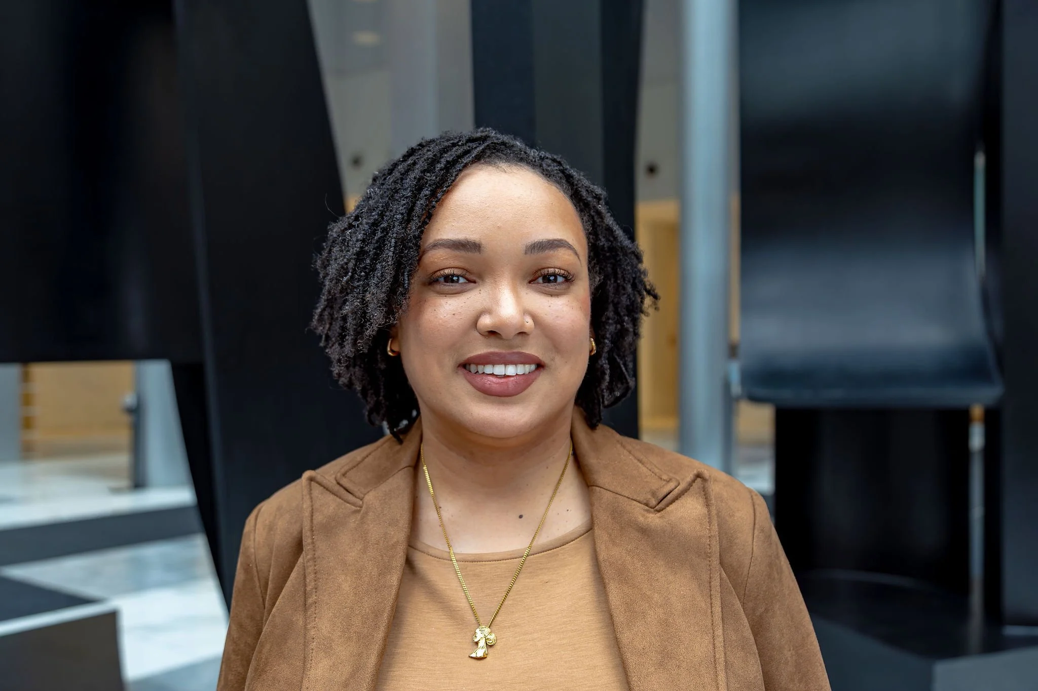 Jasmine is pictured in an open space with a textured background. She is smiling and wearing a tan blazer over a tan sweater with a gold necklace and earrings.