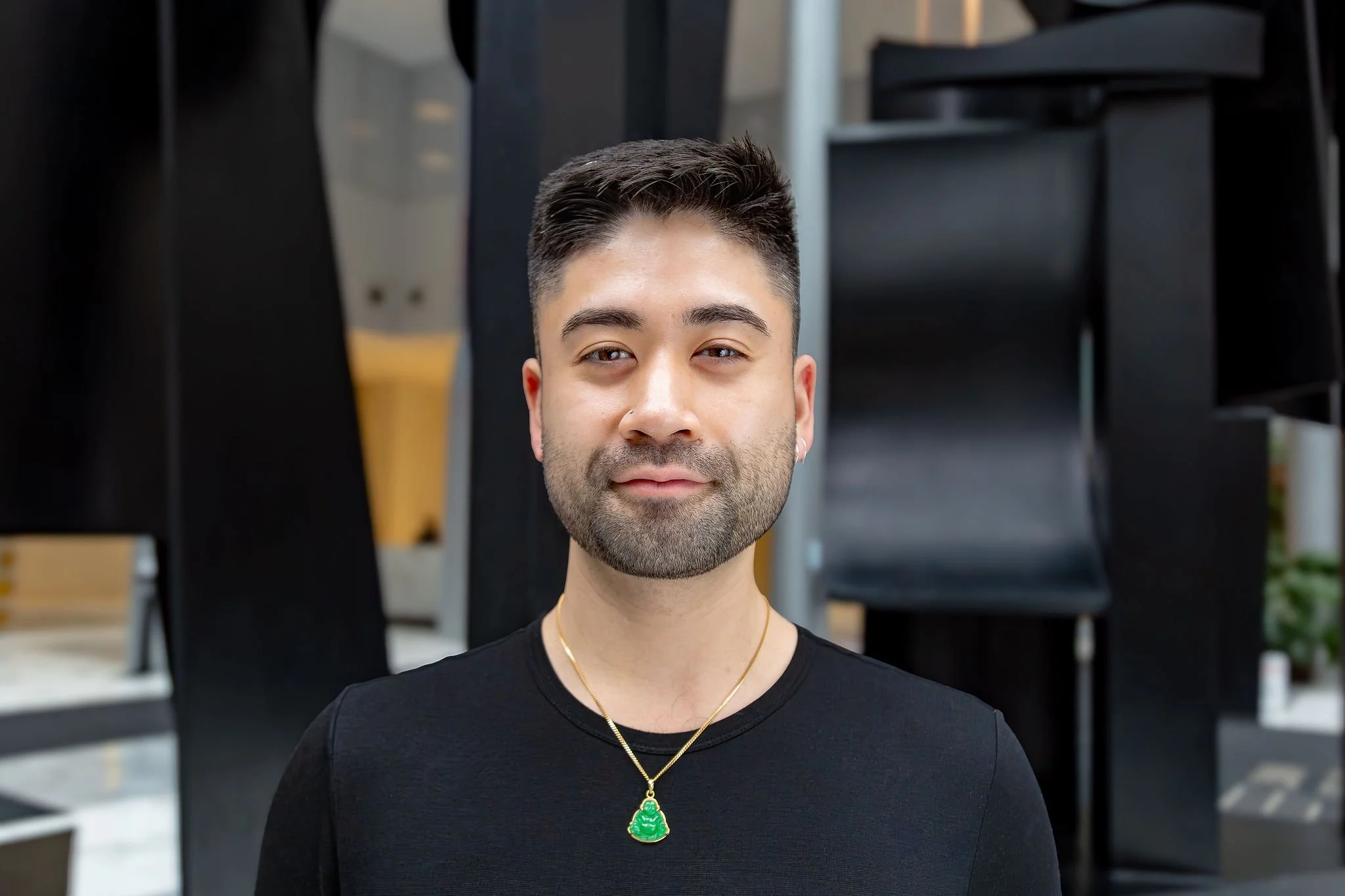 Richard is pictured in an open space with a textured background. He has a slight smile and is wearing a black shirt and a gold necklace with green pendant.