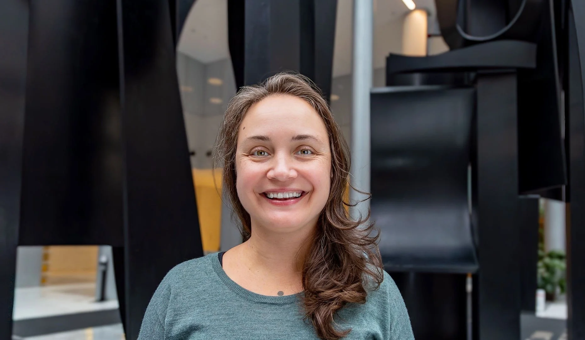 Zoe is pictured in an open space with a textured background. She is smiling and wearing a light green sweater.