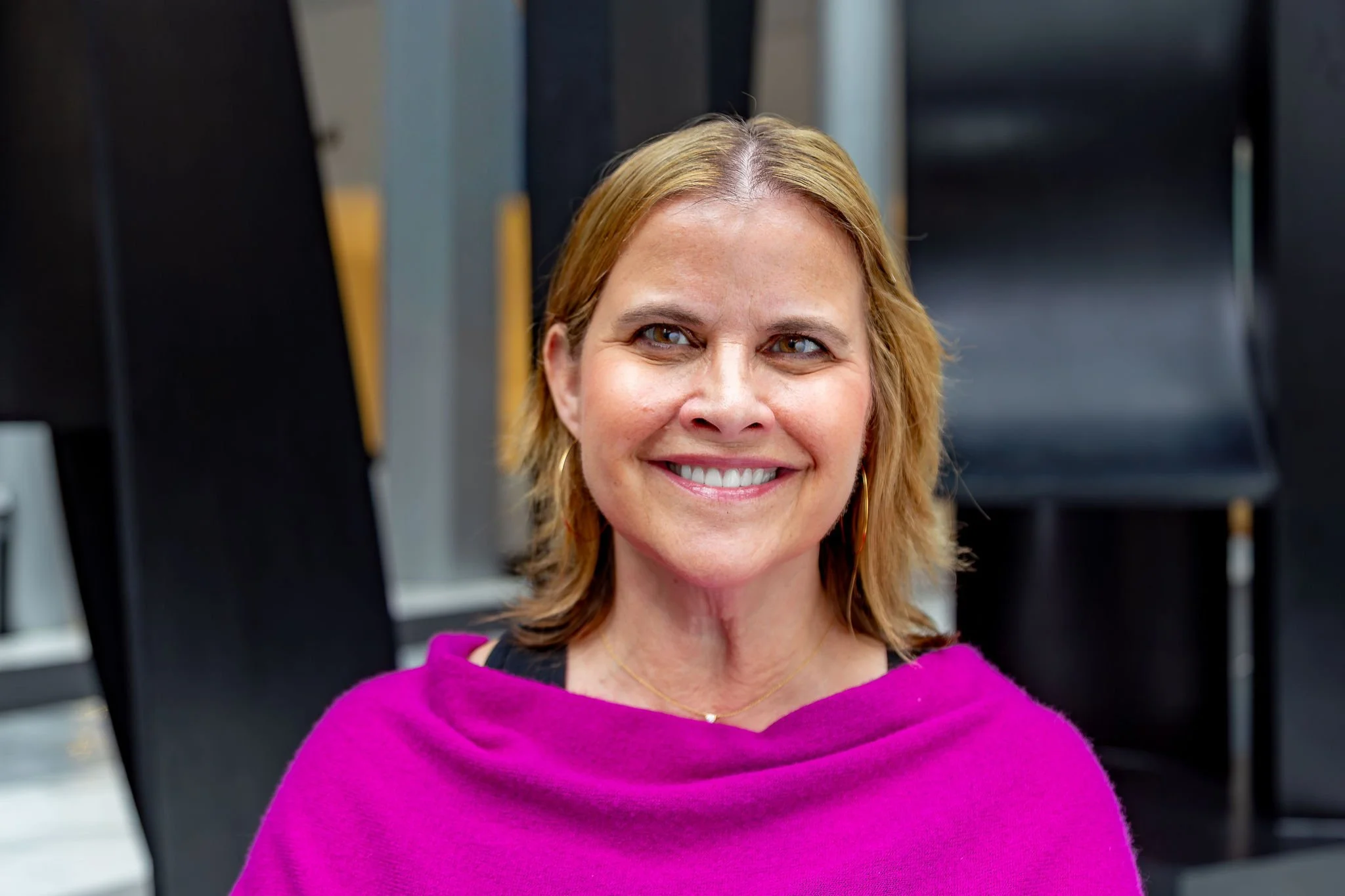 Maria is pictured in an open space with a textured background. She is smiling and wearing a magenta shawl, gold hoop earrings, and a gold necklace with a diamond pendant.