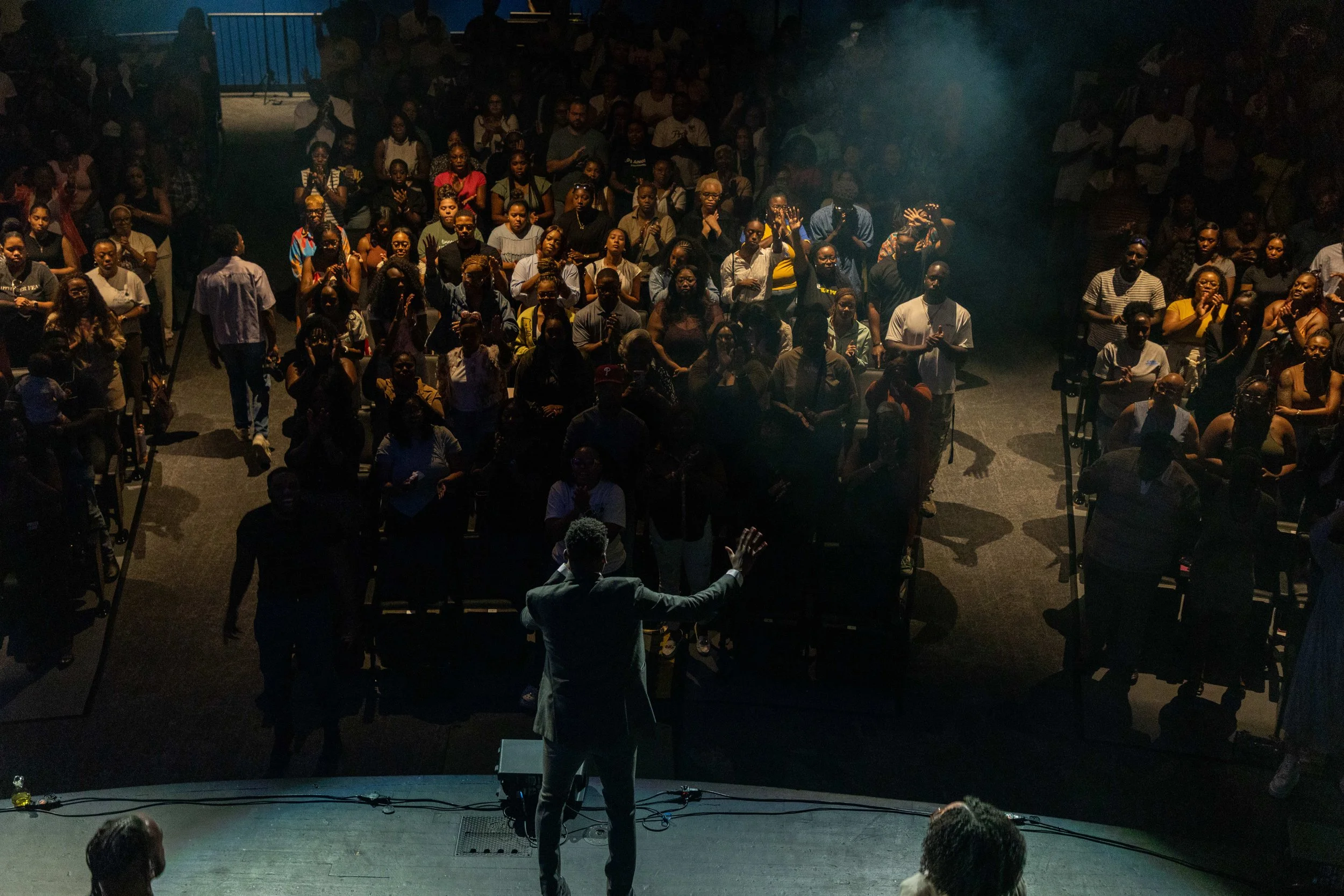 A speaker on stage addressing a large audience in an indoor venue, with dim lighting and some smoke or fog effects.