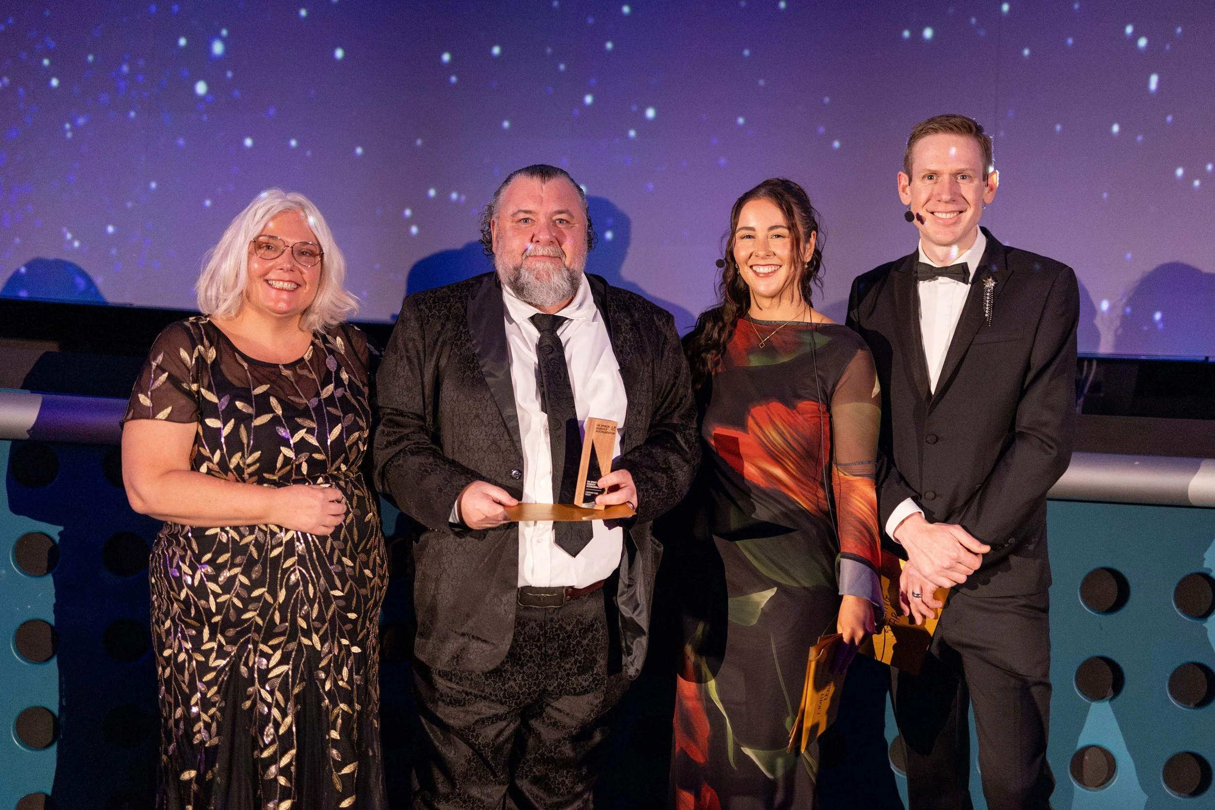 Group of four people at an awards ceremony, with two women and two men, standing on a stage with a starry background, one man holding an award.