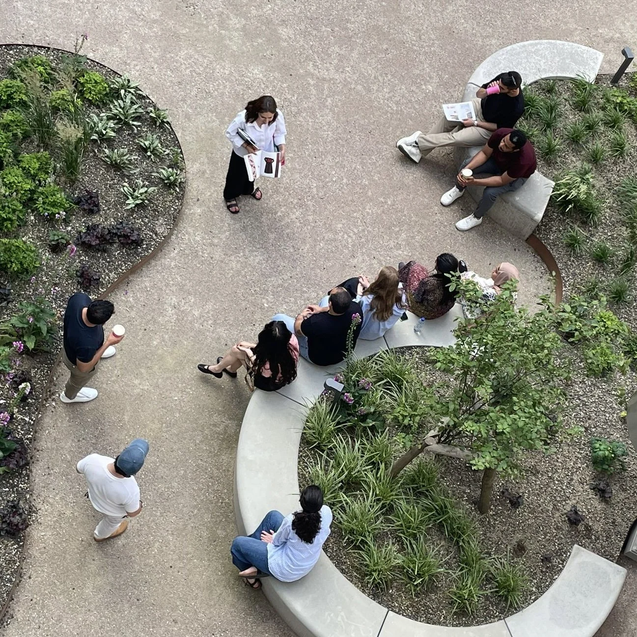 A group of people sitting and standing in an outdoor garden area, some holding coffee cups, while one woman appears to be giving a presentation or talking to the group.