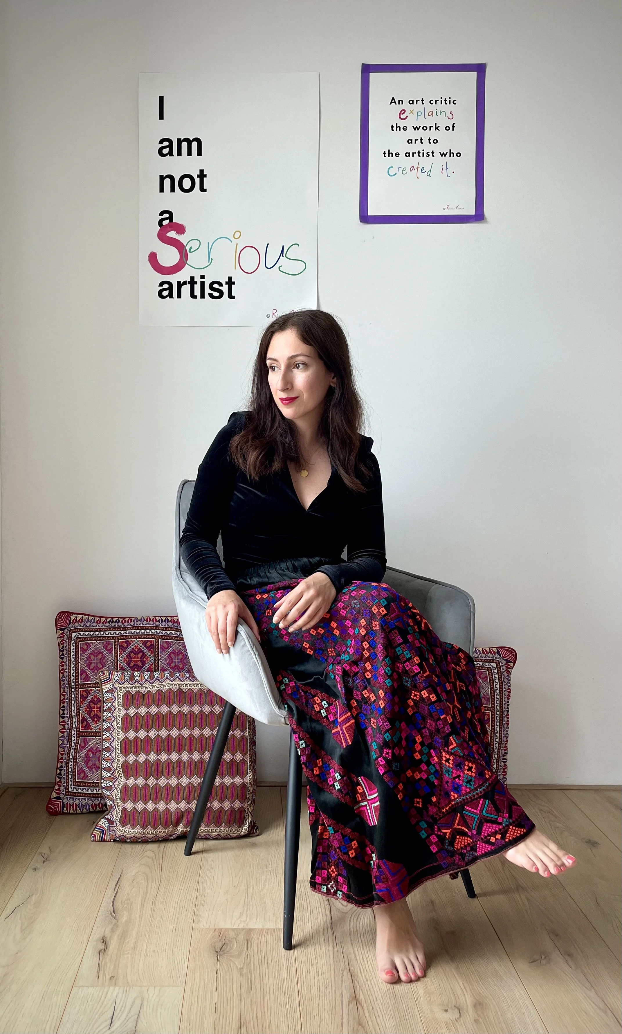 A woman with long brown hair and red lipstick sits on a gray chair with wooden legs, wearing a black top and a colorful, patterned skirt. Behind her, there are two pillows with intricate designs leaning against the wall. The wall has two posters; one with bold black and colorful writing stating "I am not a serious artist," and another framed poster with text about art critics explaining art.