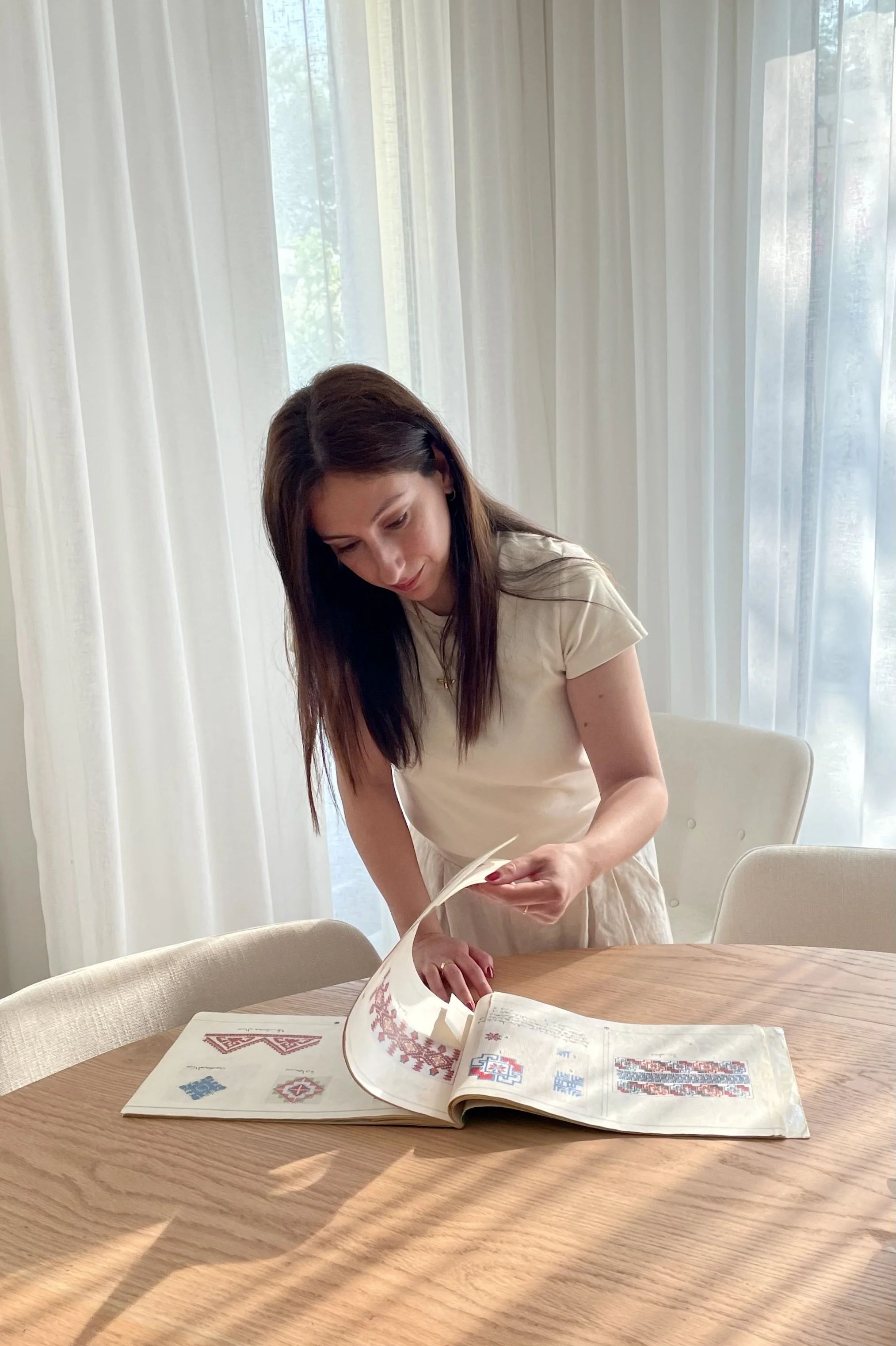 Woman looking at a book with cross-stitch patterns on a wooden table in a bright room with curtains.