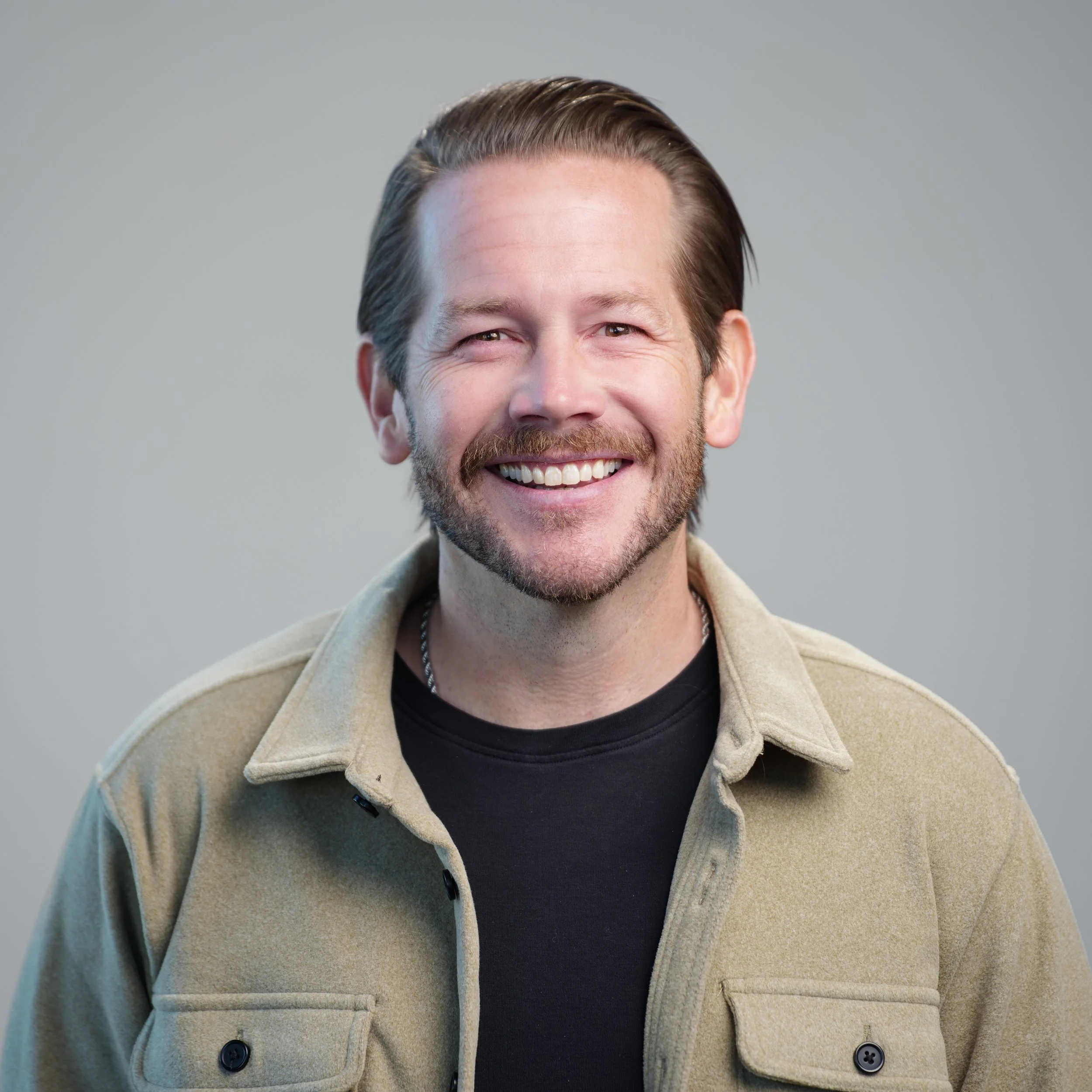 Smiling man with brown hair and beard wearing a black shirt against a gray background.