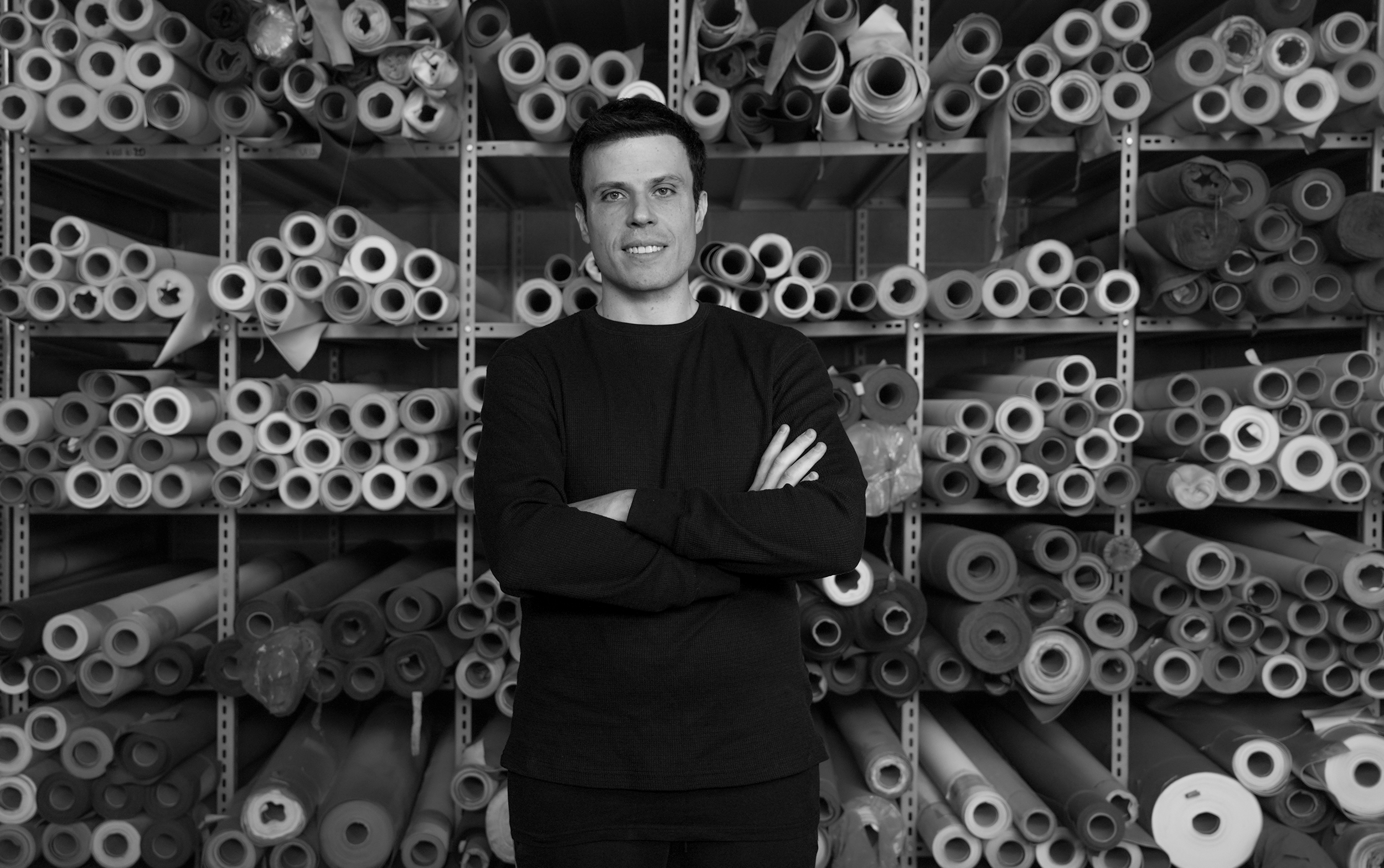 Black-and-white portrait of Vegea founder Francesco Merlino standing with crossed arms in front of shelves filled with rolled textile and material samples.