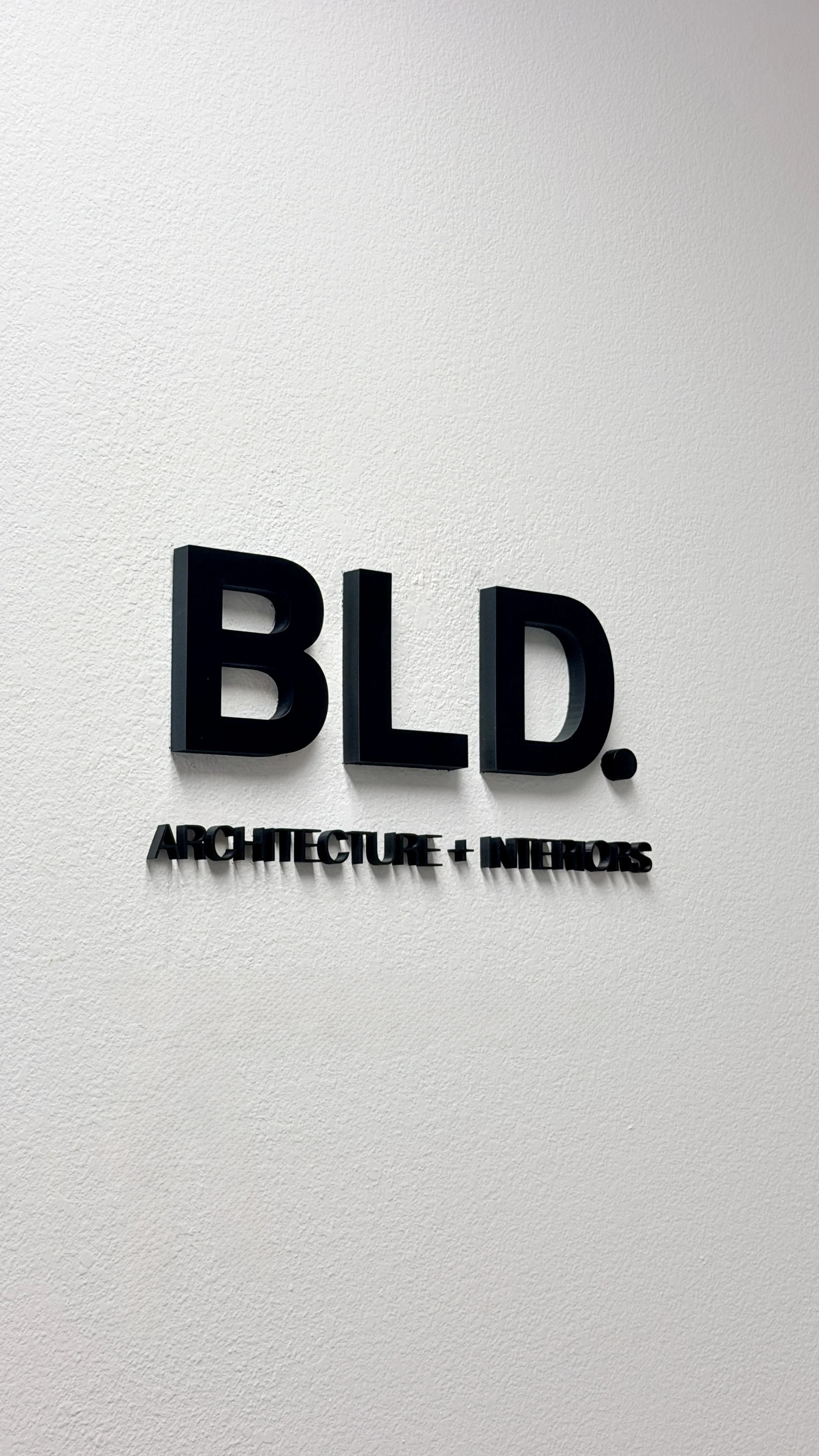 Sign with the text 'BLD.' in large black letters and 'ARCHITECTURE + INTERIORS' underneath, mounted on a white textured wall.