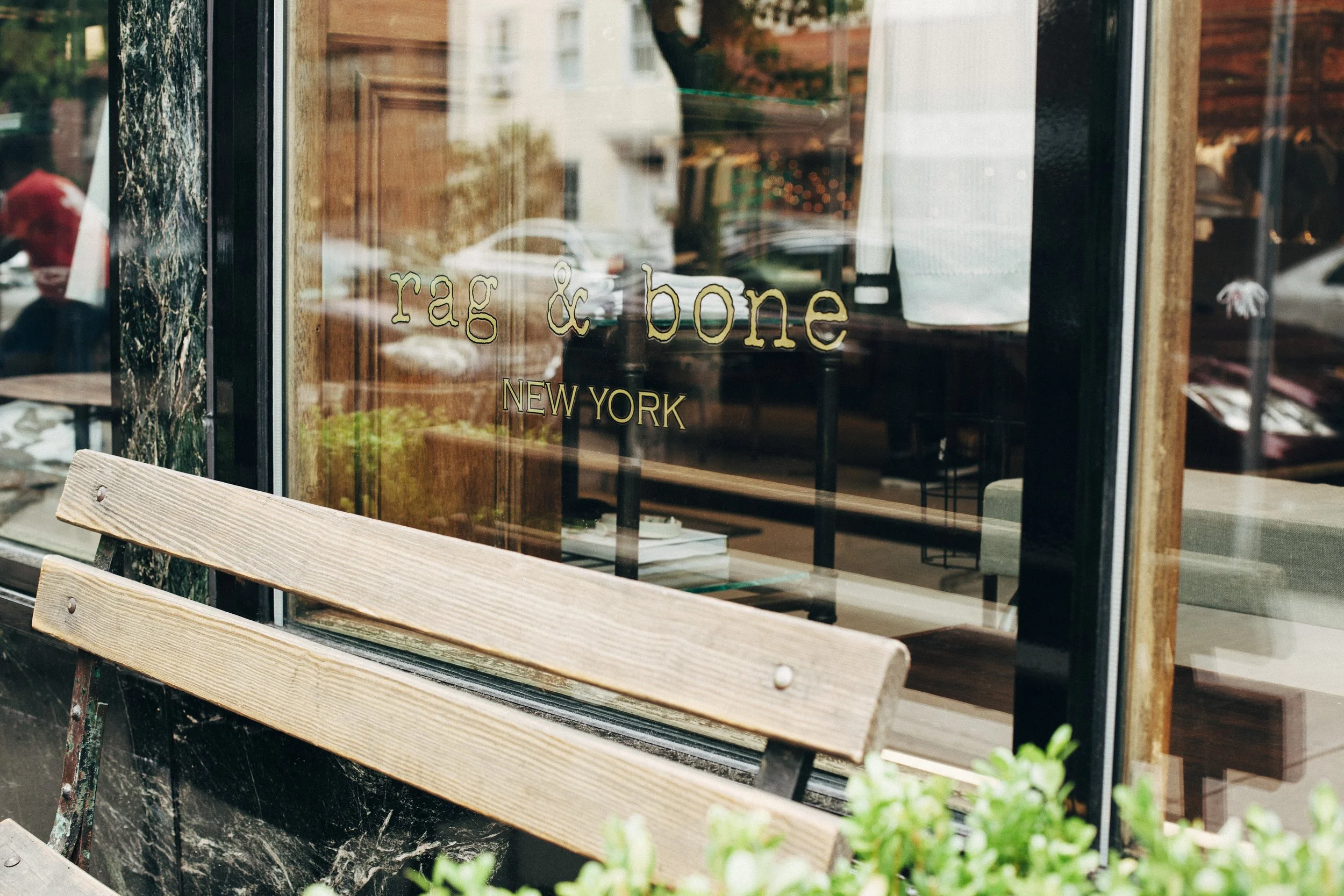 Close-up of the Rag & Bone New York storefront window in Cobble Hill, Brooklyn, featuring minimalist gold branding, a wood bench, black stone façade, and reflections of the surrounding neighborhood.