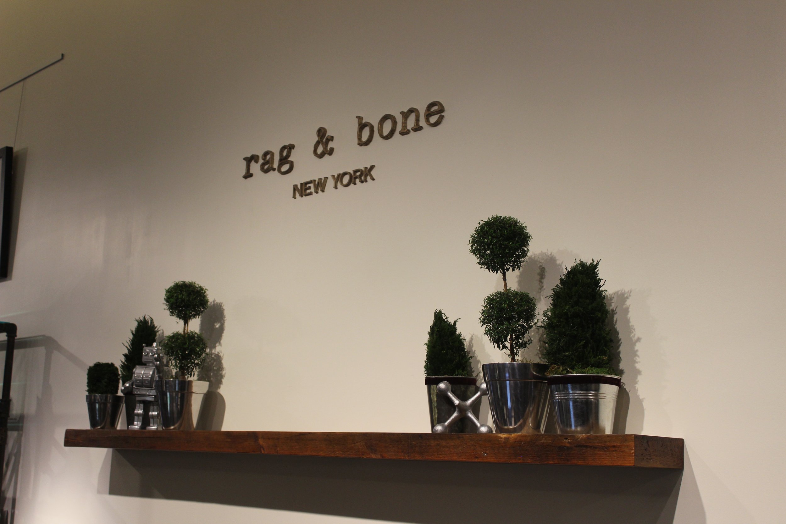 Rag & Bone Manhasset retail interior featuring minimalist Rag & Bone New York wall signage above a natural wood display shelf with sculptural topiary plants