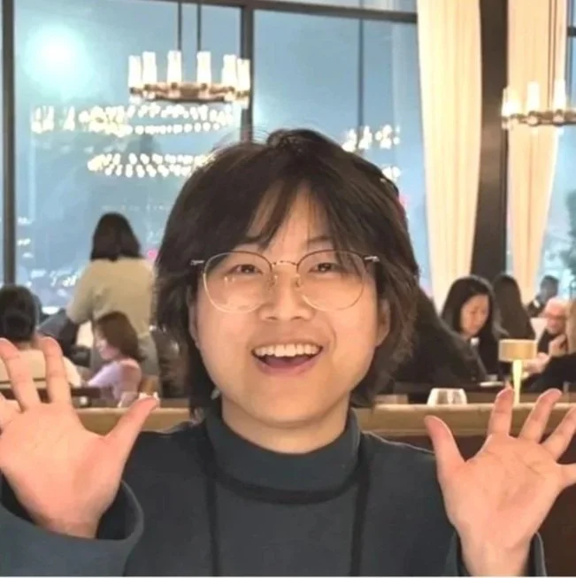 Happy woman with glasses smiling and raising hands in a restaurant with large windows and chandelier lights in background.