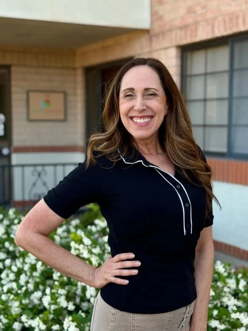 A woman smiling with glasses, long brown hair, and a floral top, standing outdoors with green foliage in the background.