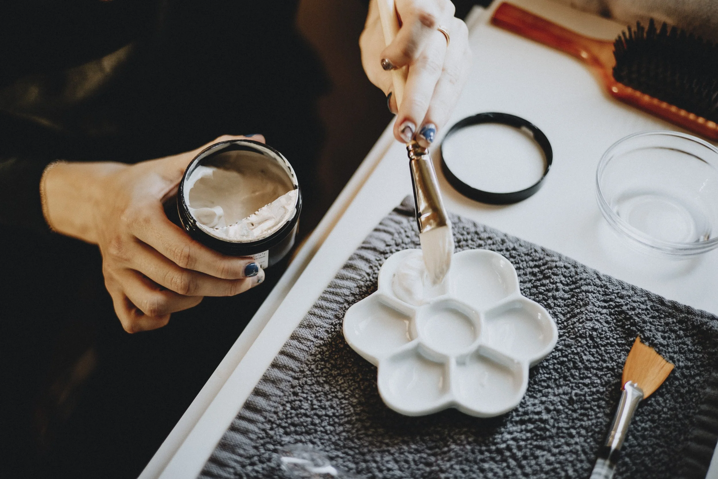 Person filling a tray with white wax using a spatula, with a container of wax and a flower-shaped mold on a textured cloth.