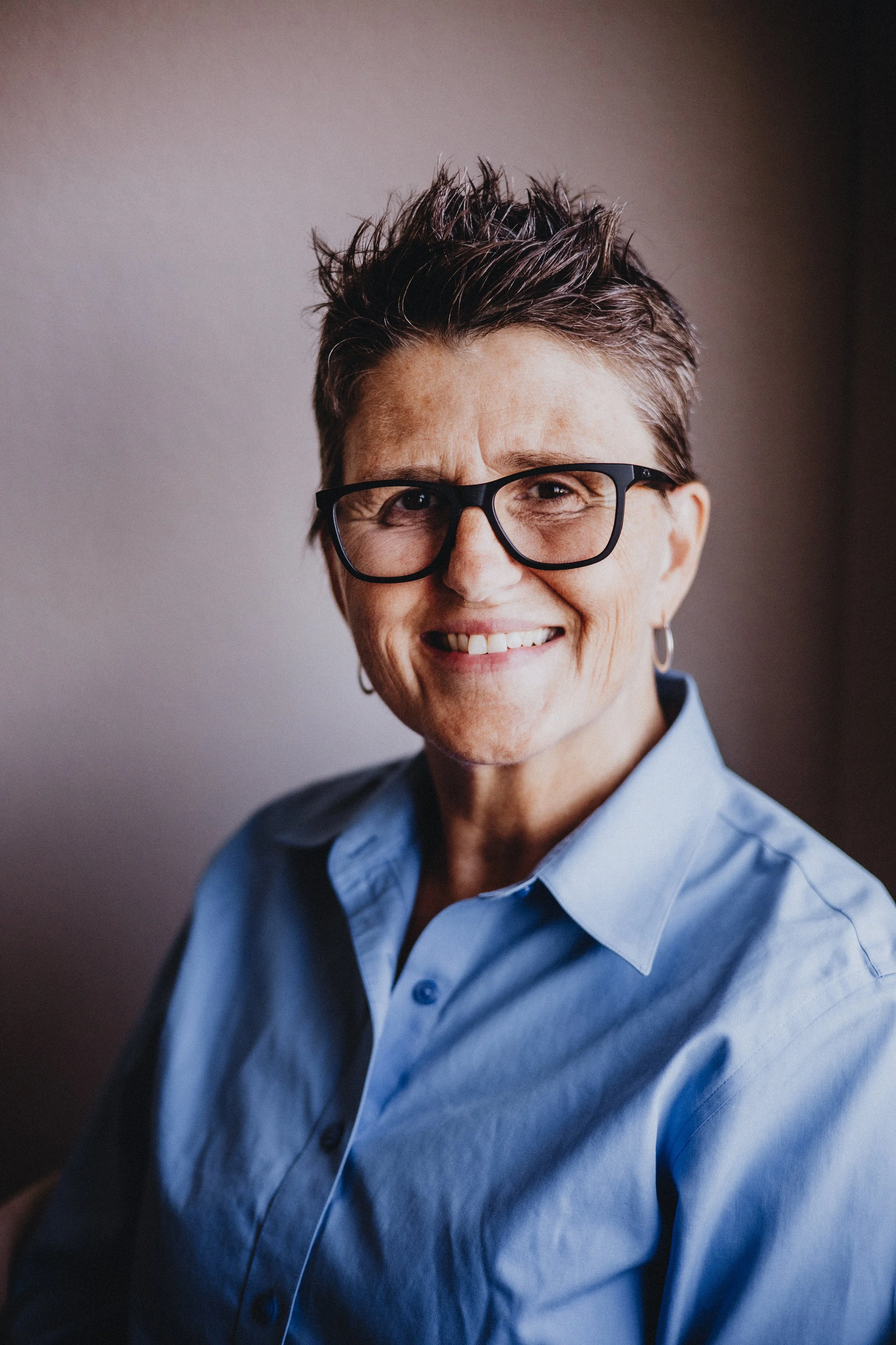 A smiling woman with short gray hair, wearing glasses, a floral blouse, layered necklaces, and a watch, sitting in front of green leafy plants.