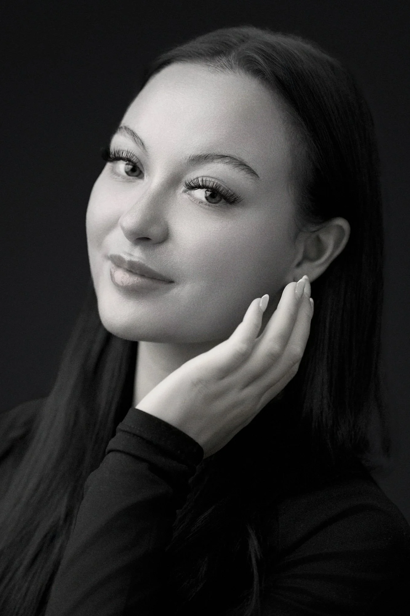 Black and white portrait of a woman with long hair, wearing a dark top and light makeup, posing with one hand near her face against a dark background.